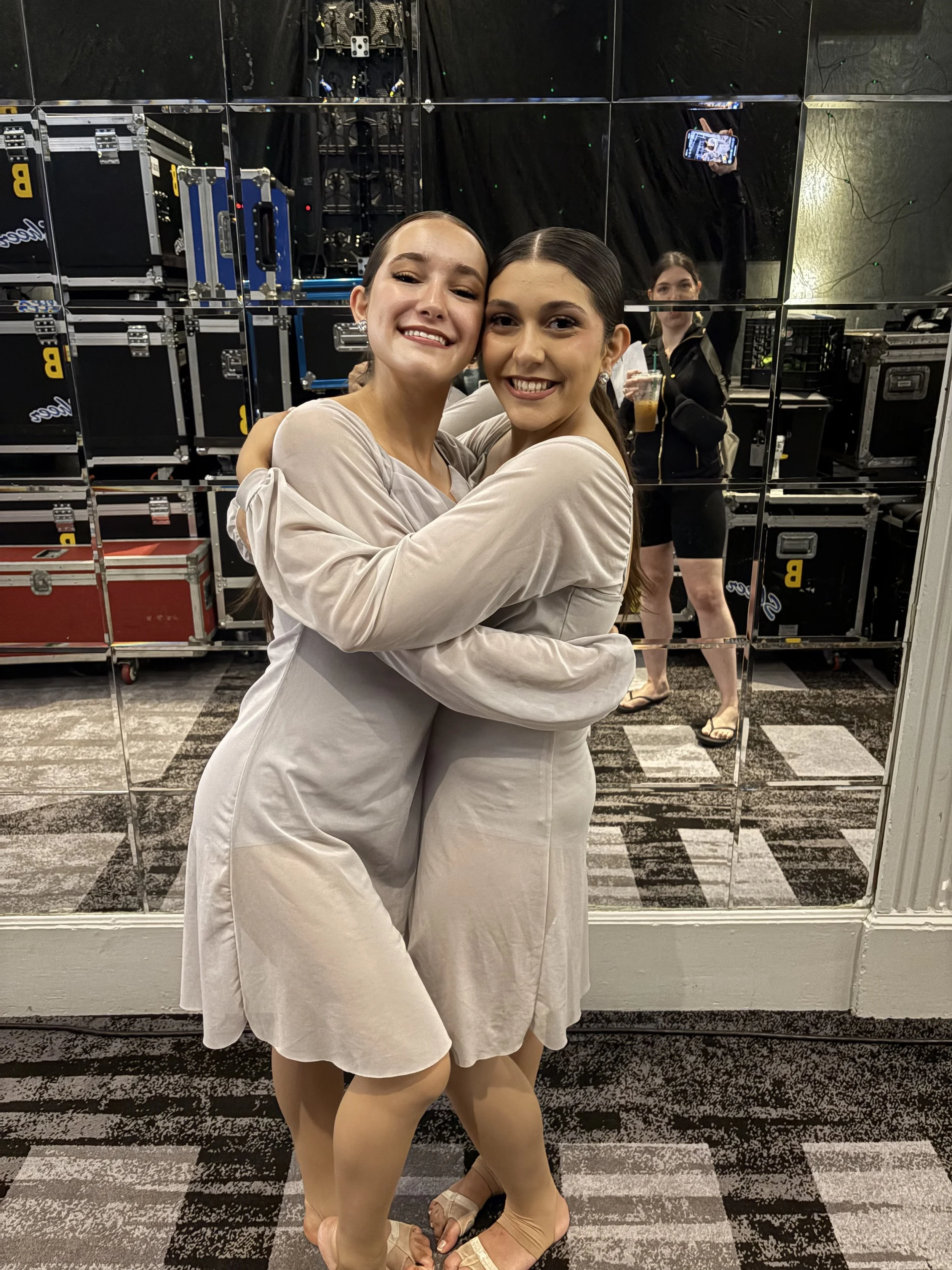 Two women smiling and hugging, wearing light-colored dresses, in front of a mirror with a dance or performance area behind them, with a third woman in the background holding a drink and taking a selfie.