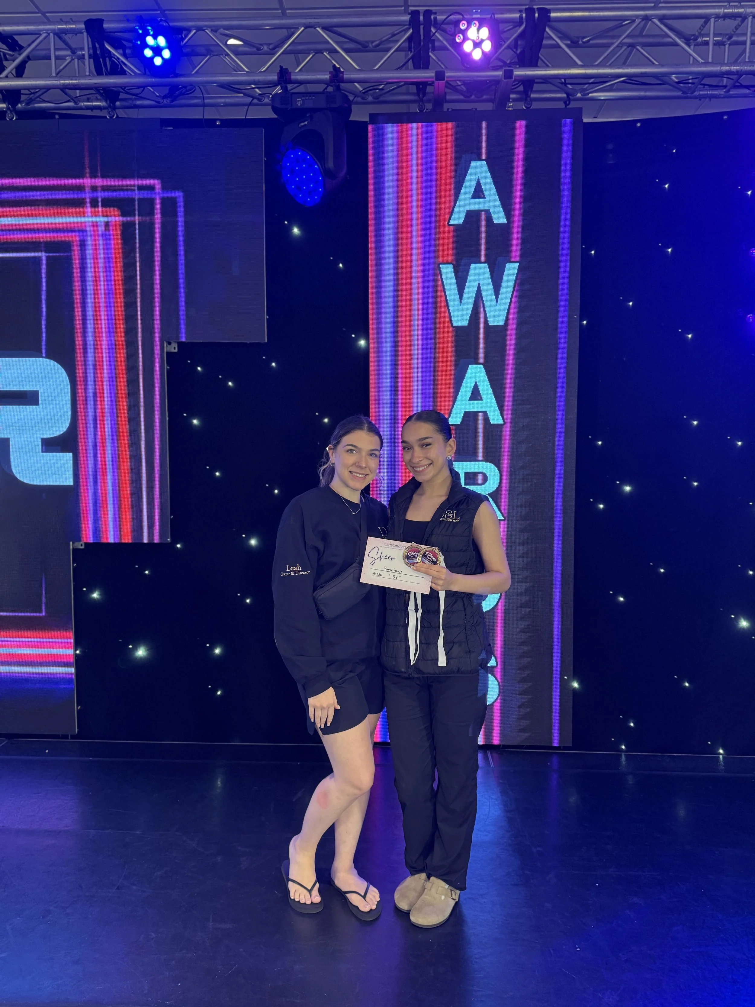 Two young women standing on a stage, one holding a certificate, with a digital sign behind them displaying the word 'AWARE'.