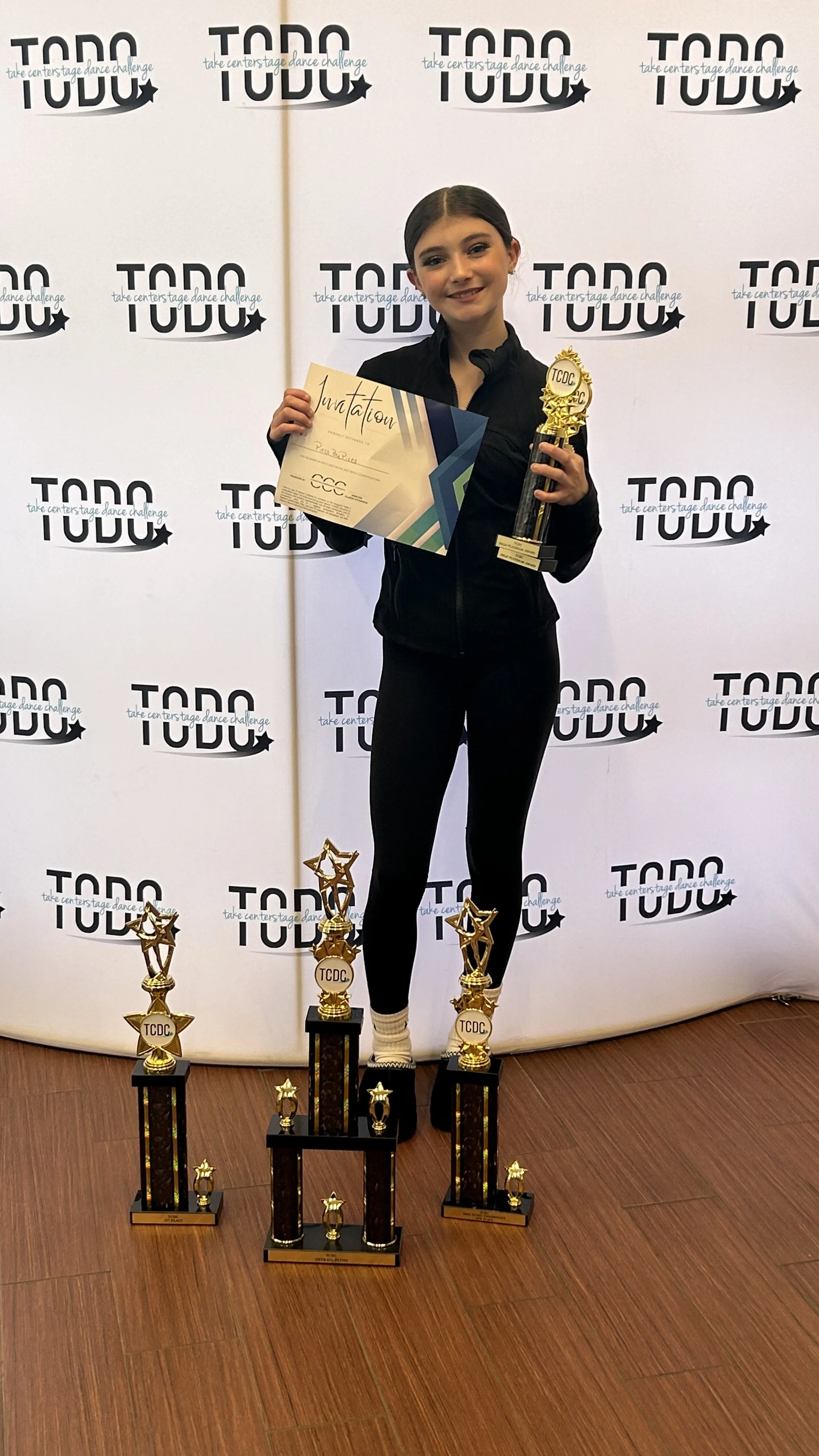 Young female dancer standing on a poster with the words 'TADC' and 'take center stage dance challenge' repeating in the background. She is holding a certificate and a trophy, surrounded by multiple gold trophies on the floor.