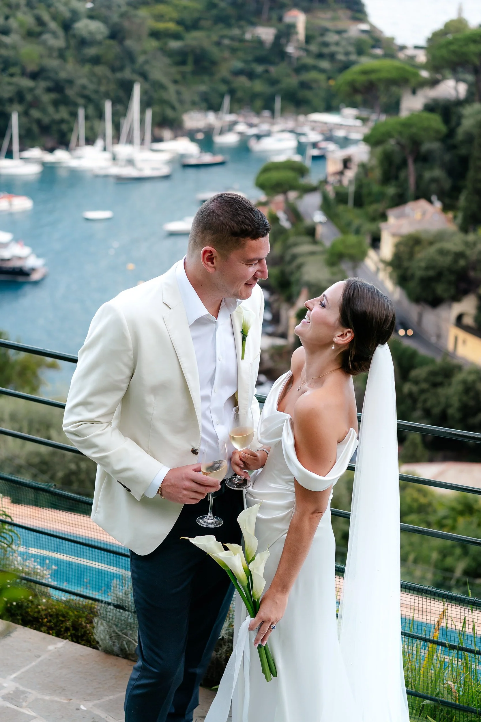 A bride and groom smiling at each other with a scenic Portofino harbour, holding champagne glasses,bride holding a bouquet of calla lilies Portofino at a Belmond Splendido Wedding. Photos taken by editorial destination wedding photographer