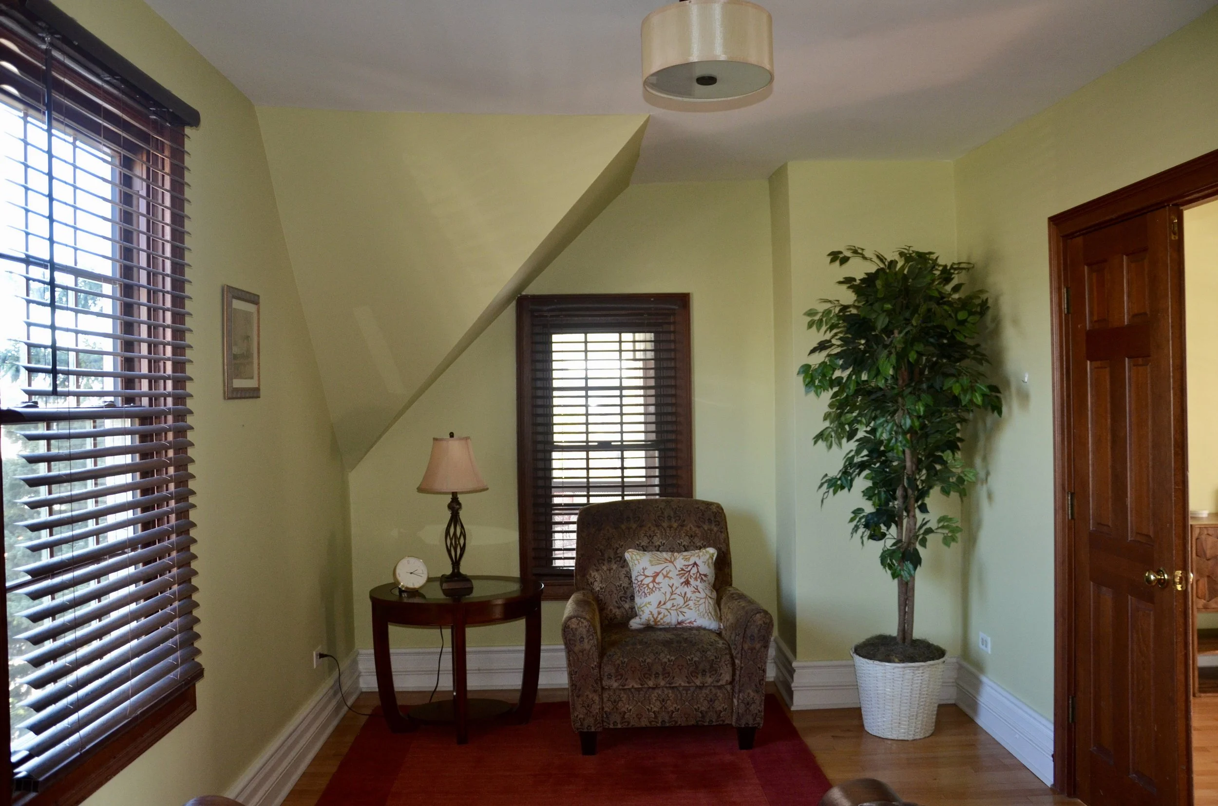 Living room corner with green walls, a window with wooden blinds, a patterned armchair with a decorative pillow, a small side table with a lamp and clock, a tall potted tree, and a partially open wooden door.