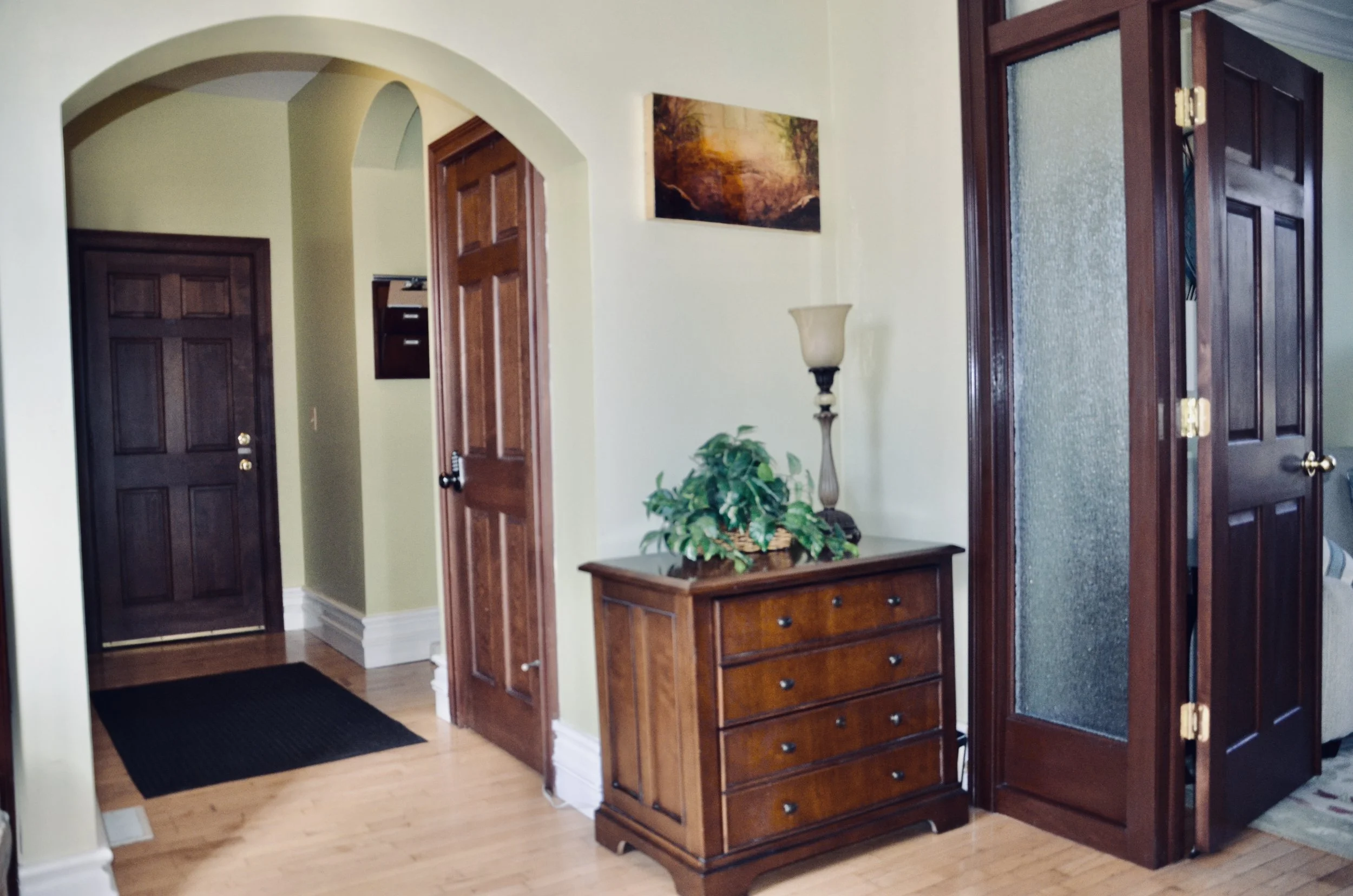 Living room with wooden doors, a small wooden dresser with a plant and a lamp on top, a framed landscape painting on the wall, and an archway leading to a hallway with a black doormat.