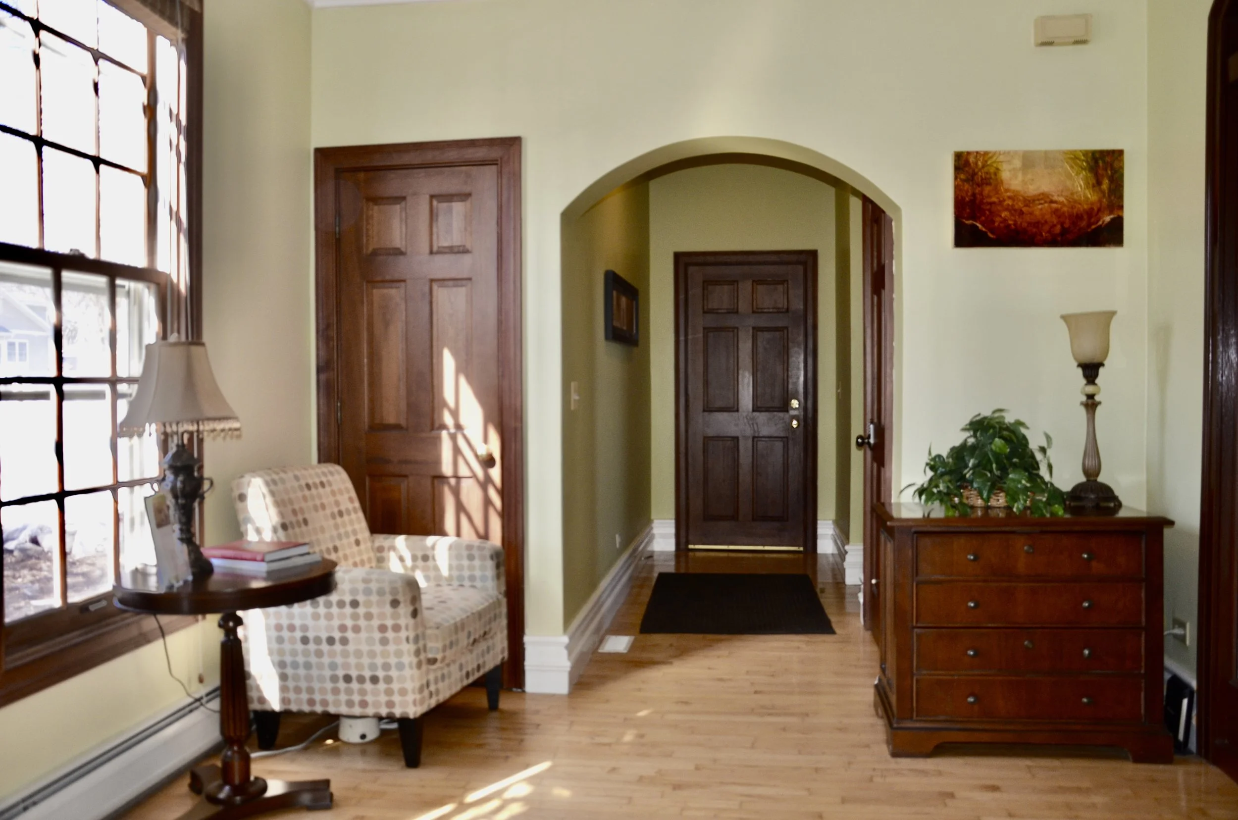 View of a home interior with a front door, a window with divided panes, a checkered armchair, a side table with books and a lamp, a wooden dresser with a plant and a lamp, and decorative wall art.