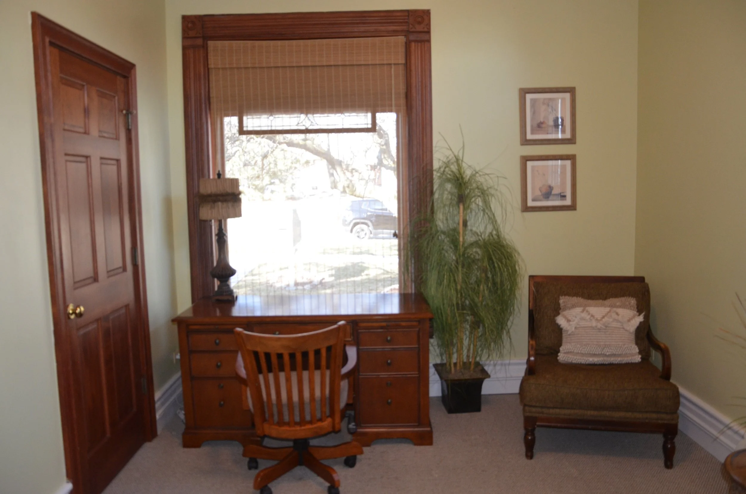 Home office with wooden desk and chair near a window, green wall, potted plant, framed artwork, and a wooden chair with cushion.