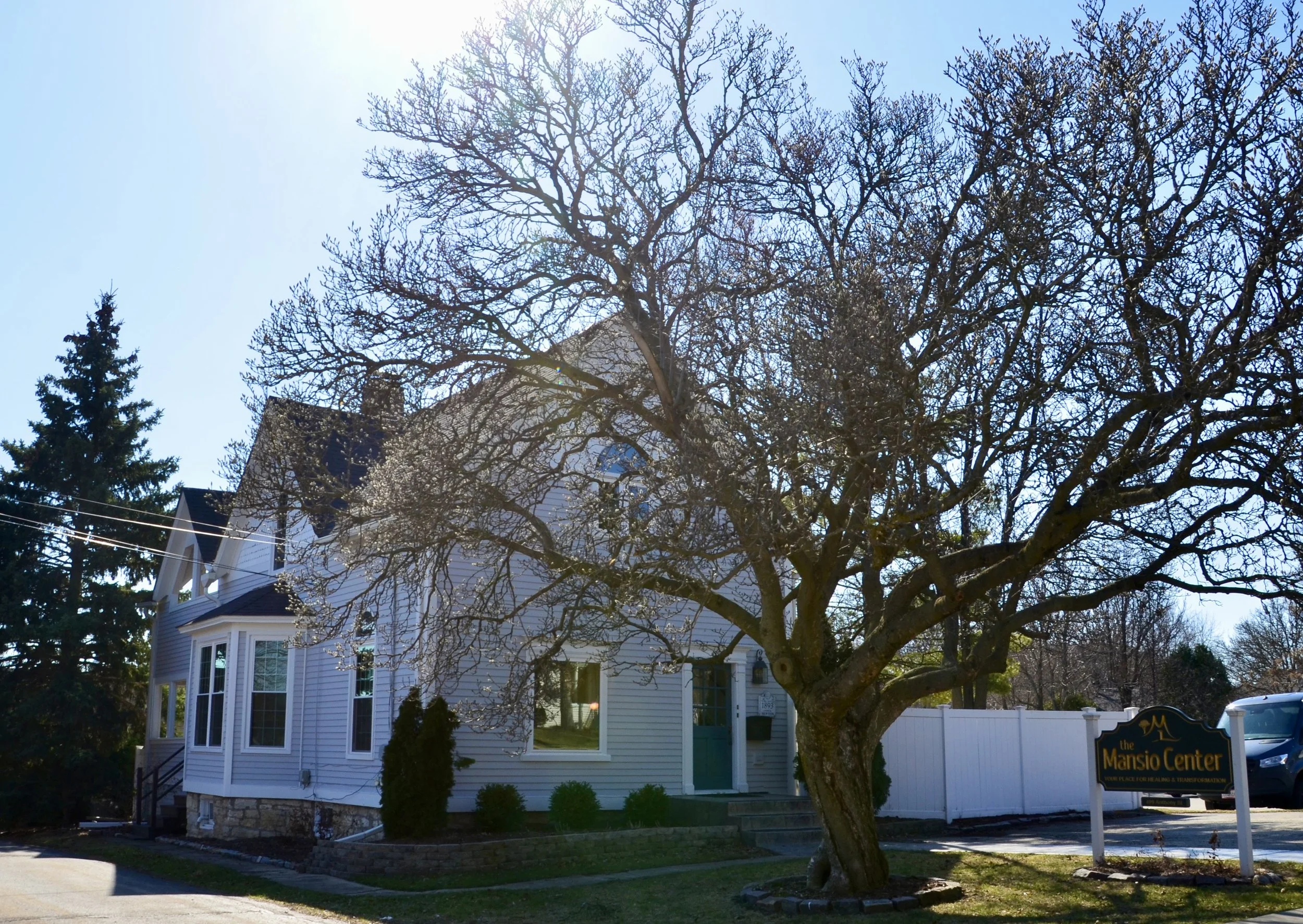 A white house with a large, leafless tree in front, under a bright blue sky, with a sign reading "the Manso Center" nearby.