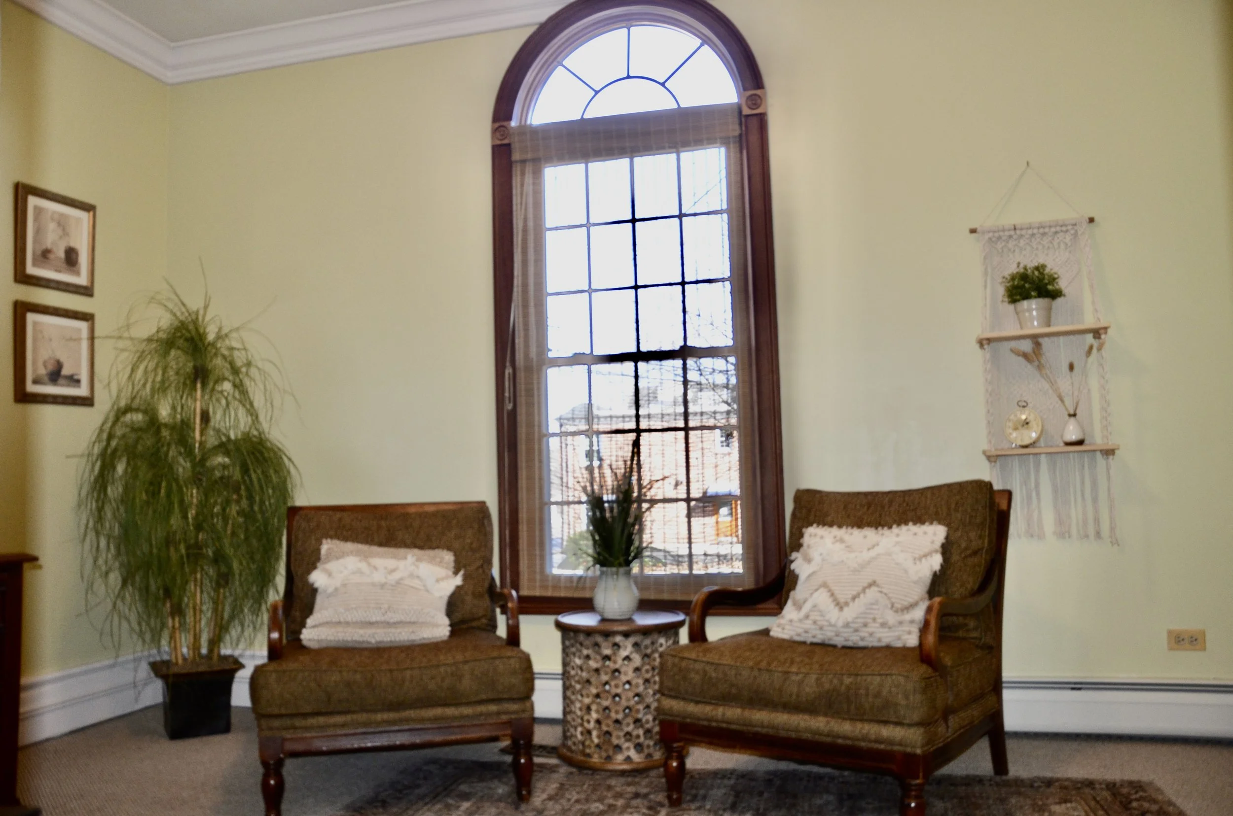 Living room with two vintage armchairs, a small round side table with a potted plant, a large window with wooden framing, an indoor plant,-framed pictures, and decorative wall shelves with plants and vases.