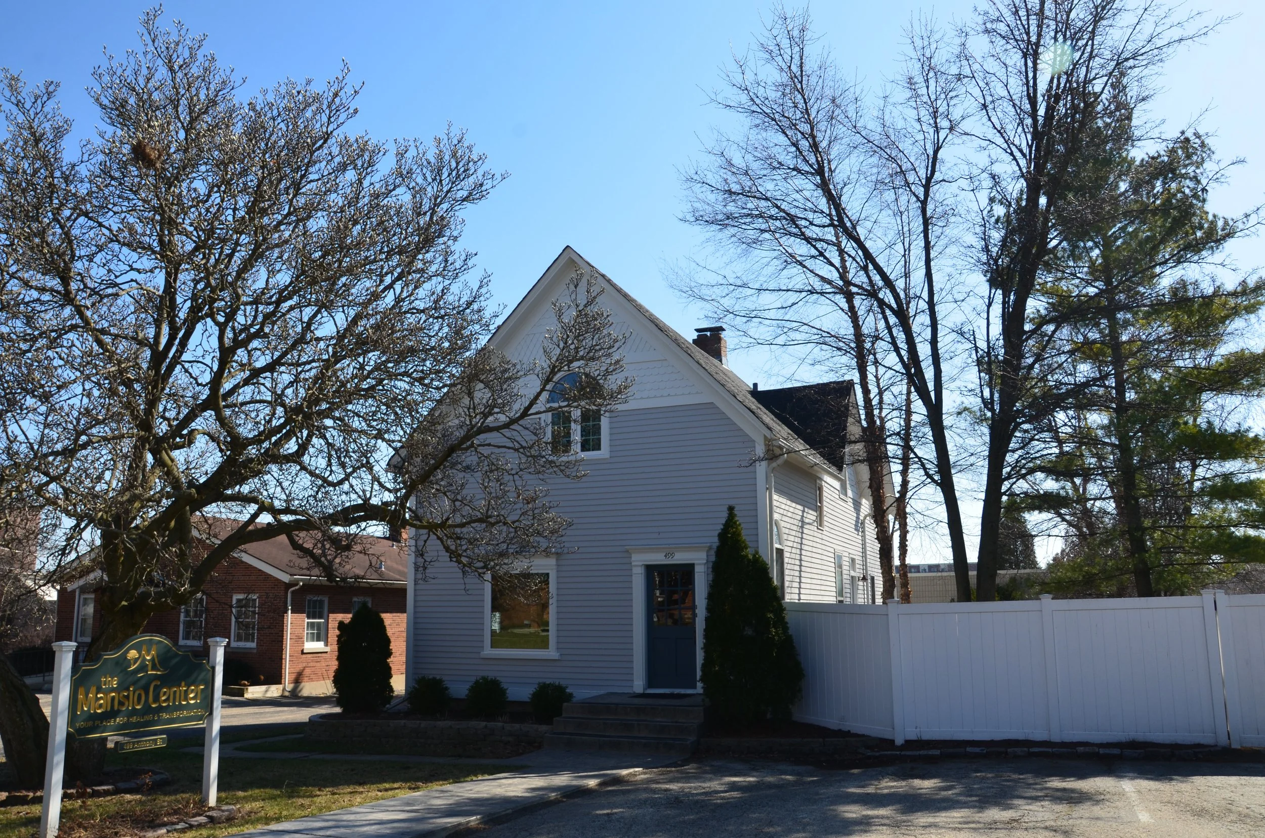A white house with a gabled roof and a small front porch, surrounded by leafless trees and a white fence, with a sign that reads 'The Mansio Center' on the left side of the image.