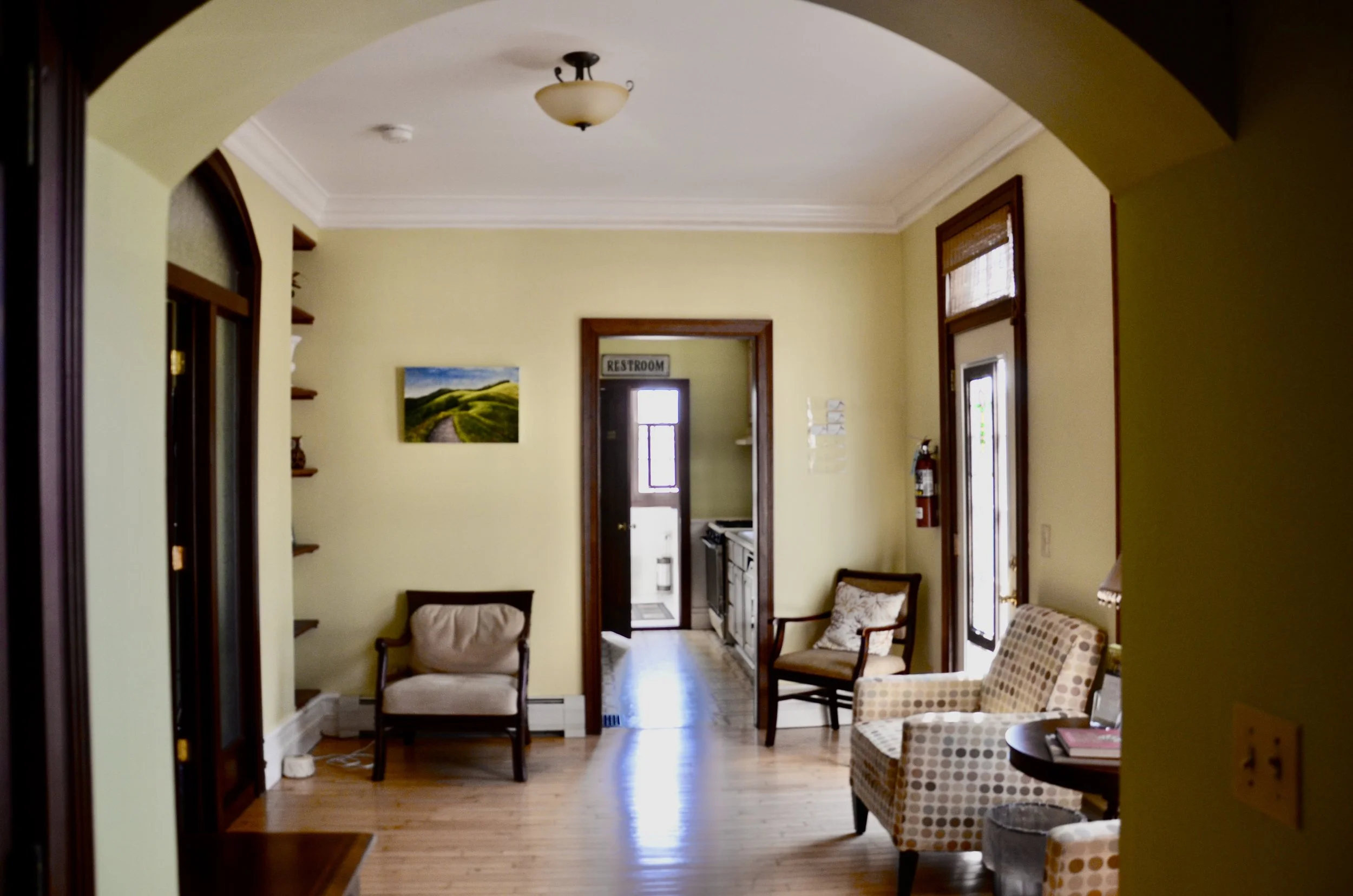 Living room with yellow walls, wooden flooring, armchairs, a bookshelf, a window, and an open door leading to a kitchen area.