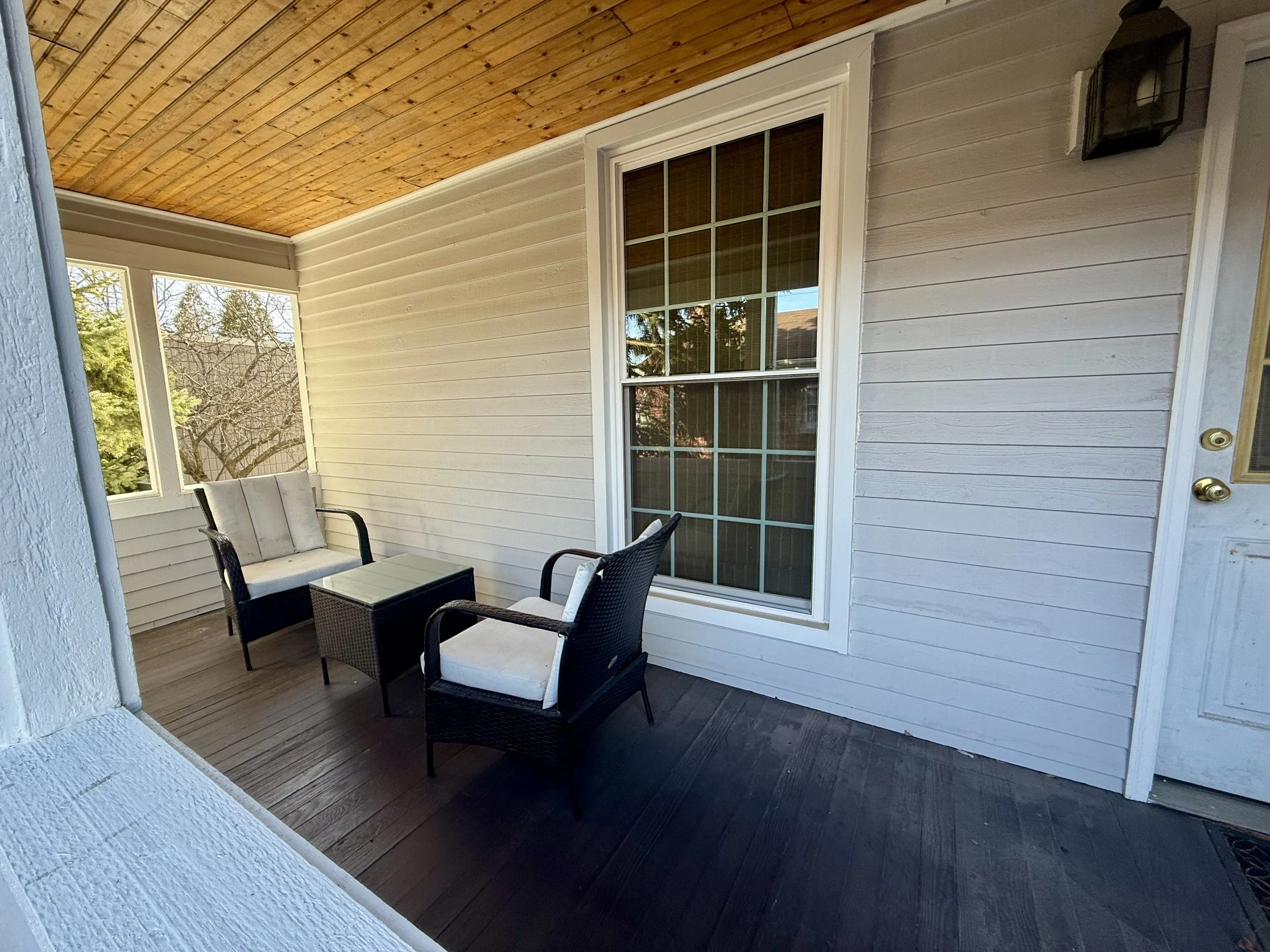 Covered porch with white siding, large window, black wicker furniture with cushions, and a small table.