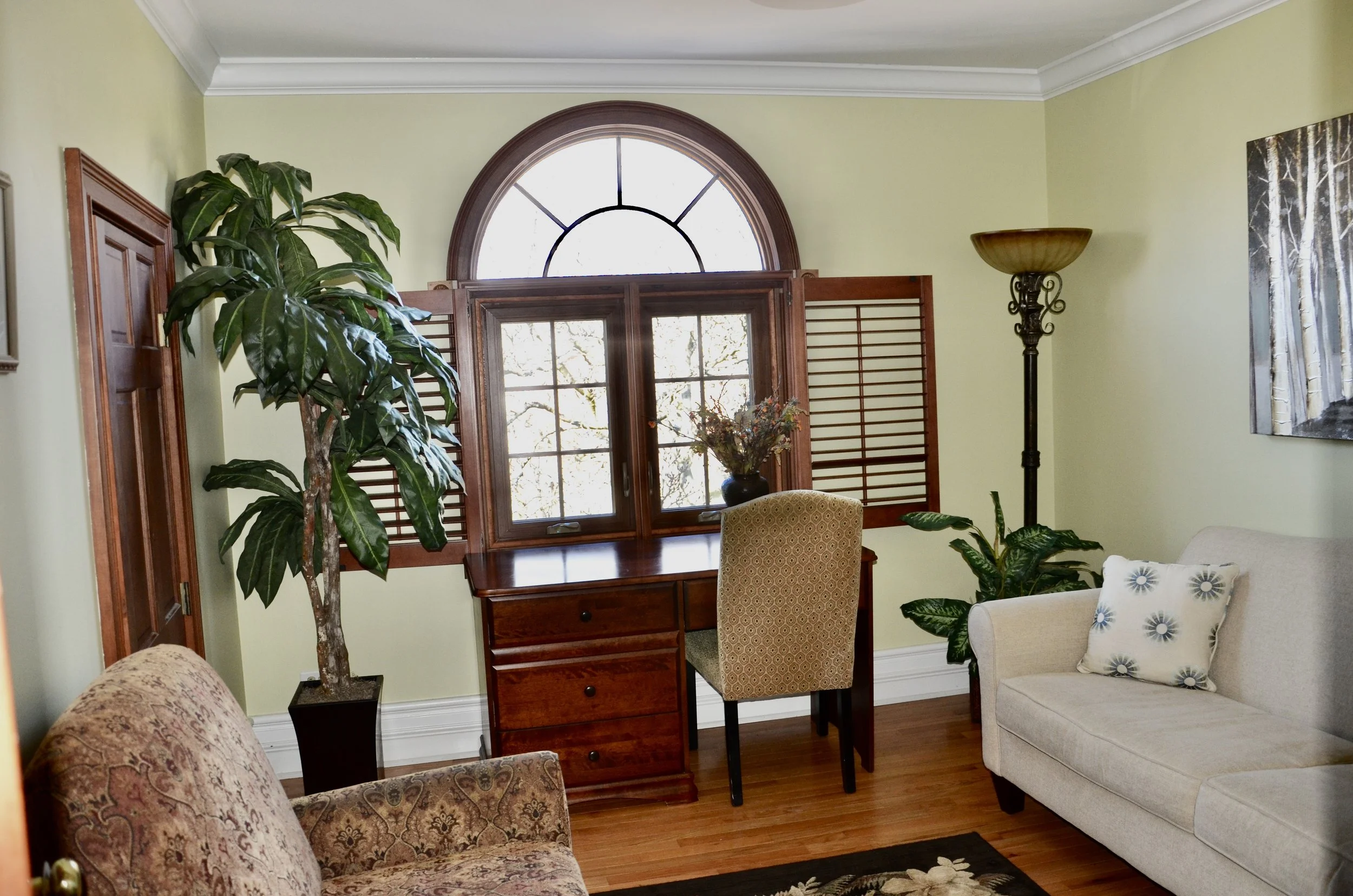 Living room with green walls, hardwood floor, large window with wooden shutters, a desk with a chair in front of the window, a tall floor lamp, a large potted plant, a beige armchair, a white sofa with a floral pillow, and framed artwork on the wall.
