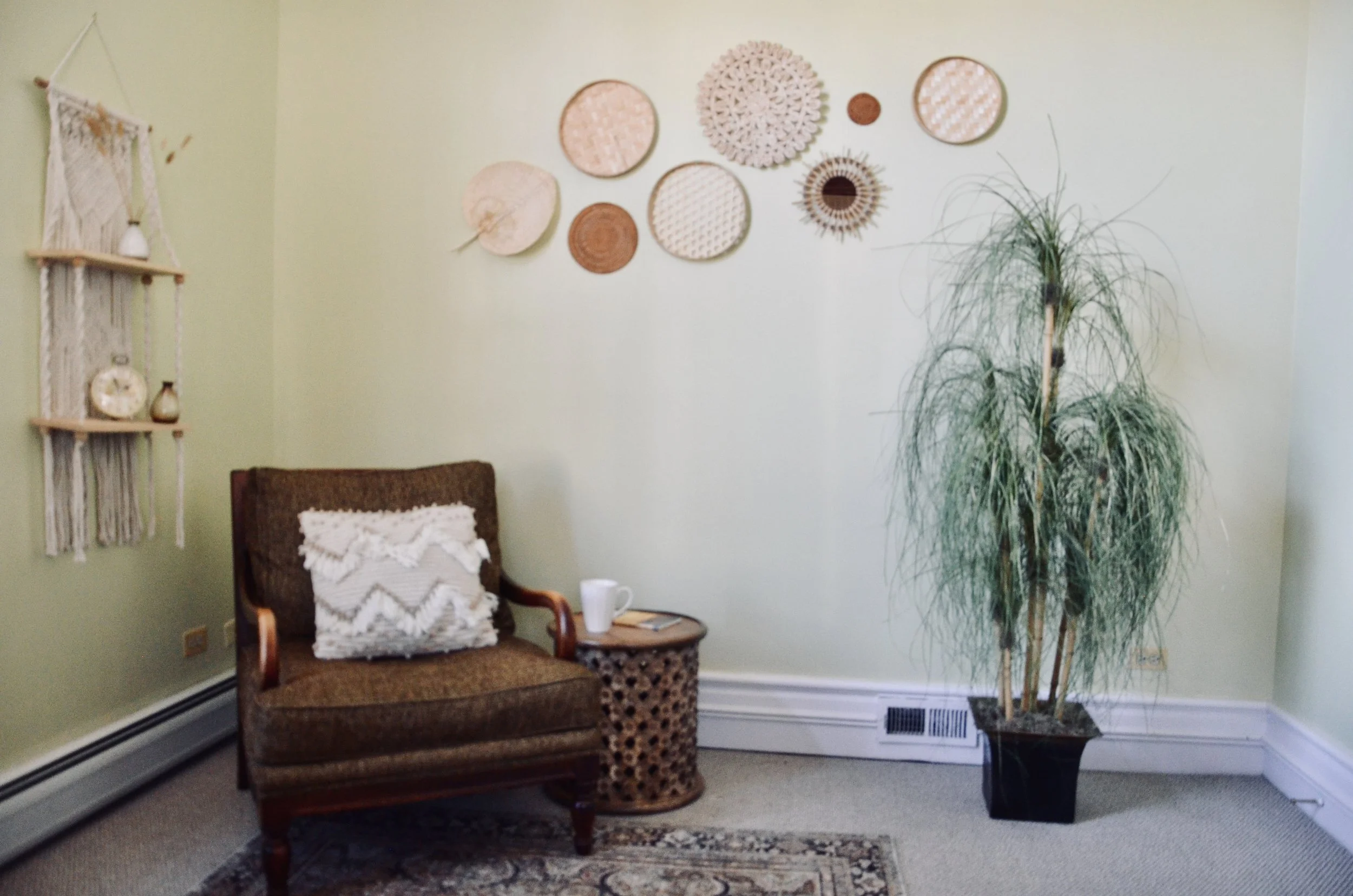 A cozy corner featuring a brown upholstered armchair with a decorative white pillow, a small round side table with a white mug and a book, a tall potted plant, and a wall decorated with a variety of woven and macramé wall hangings.