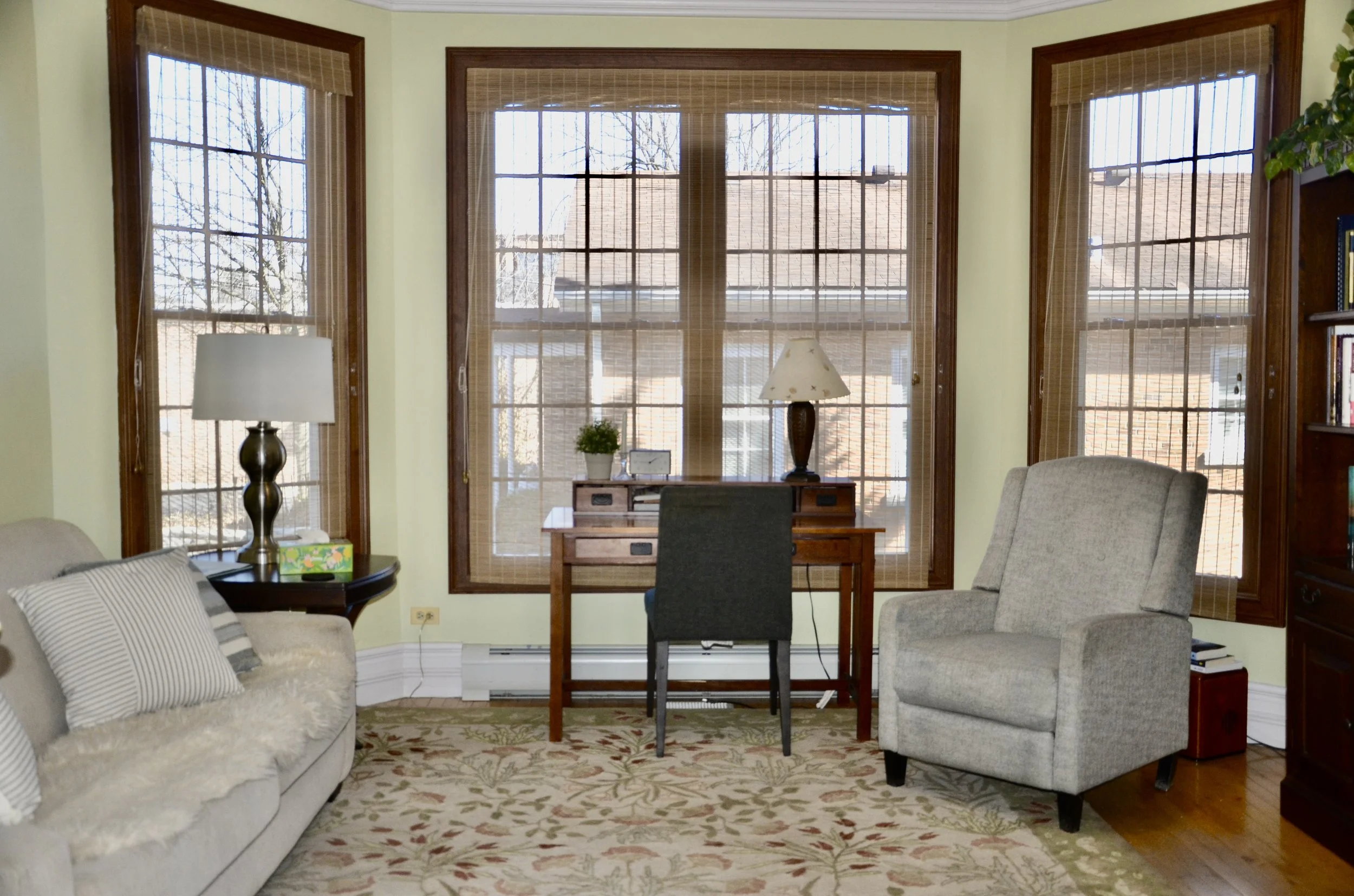 Living room with large windows, beige sofa, armchair, wooden desk with a lamp and plant, side table with a lamp, and a bookshelf.
