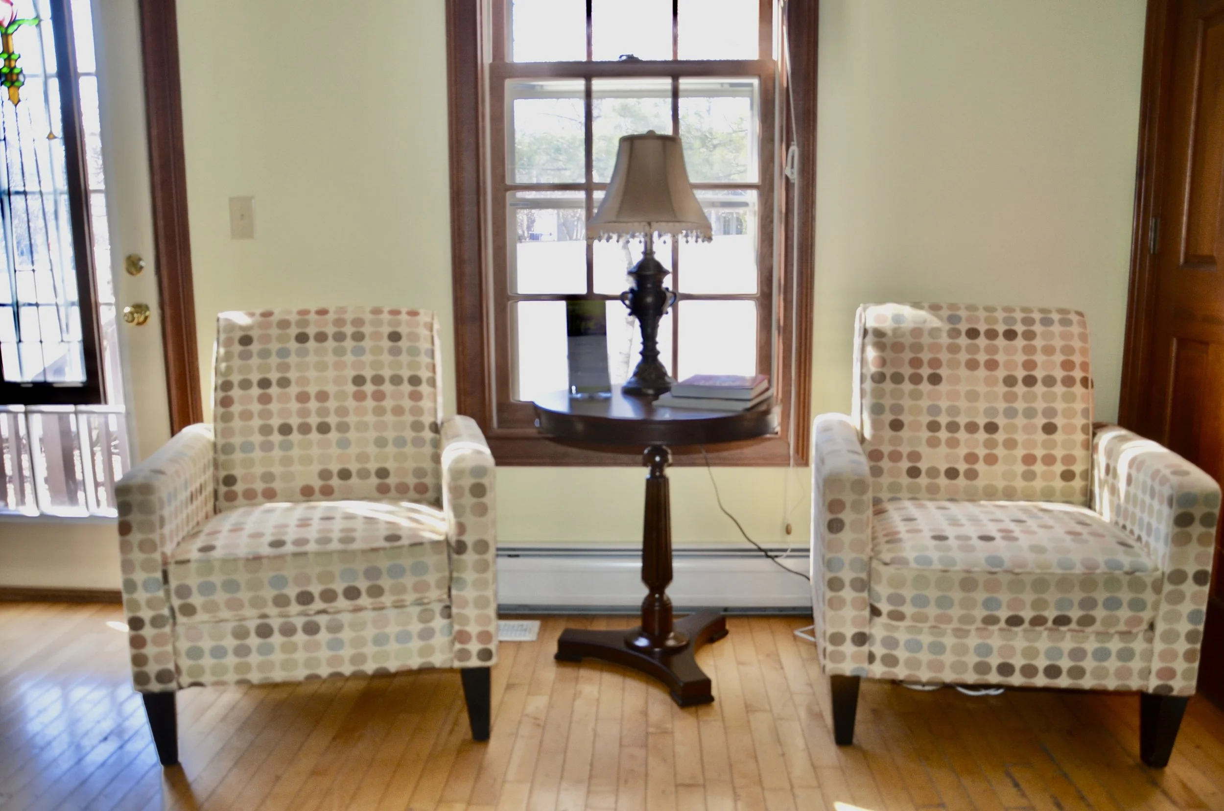 Two armchairs with polka-dot fabric upholstery sitting on a wooden floor with a round wooden side table and a table lamp between them near a window.