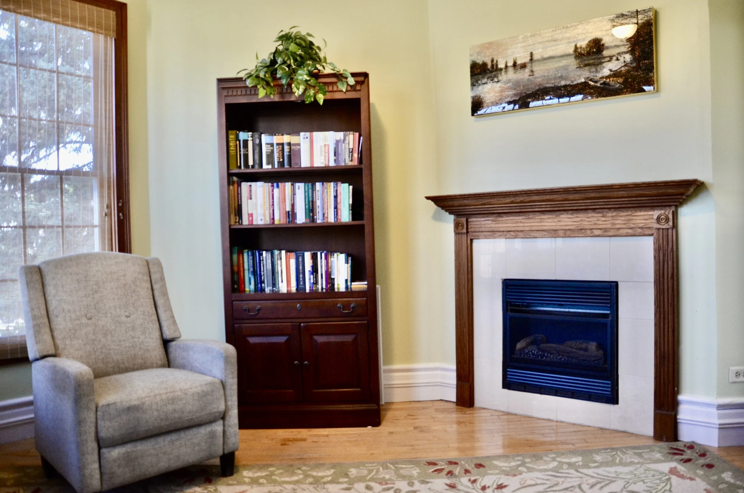 Living room with beige armchair, wooden bookcase filled with books, fireplace with wooden mantle, wall-mounted landscape painting, window with blinds, and a floral area rug.