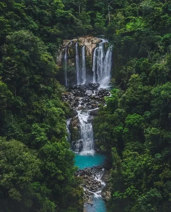 Multi-tiered waterfall flowing into a turquoise pool in a lush green forest.