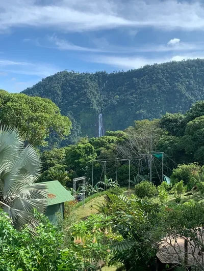 Lush green forested hillside with a waterfall, blue sky, and some structures and trees in the foreground