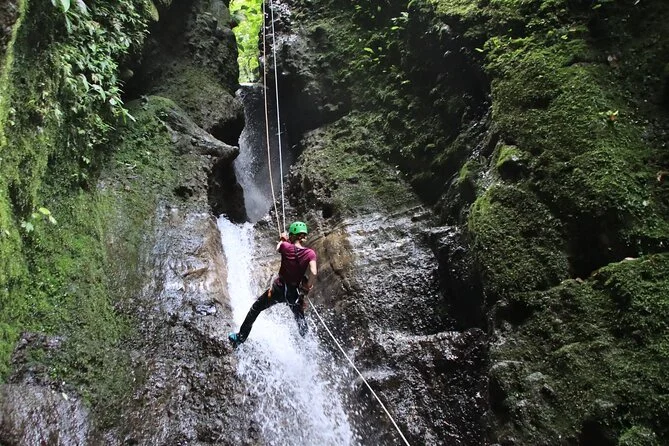 Person wearing a green helmet and maroon shirt rappelling down a mossy, rocky waterfall.