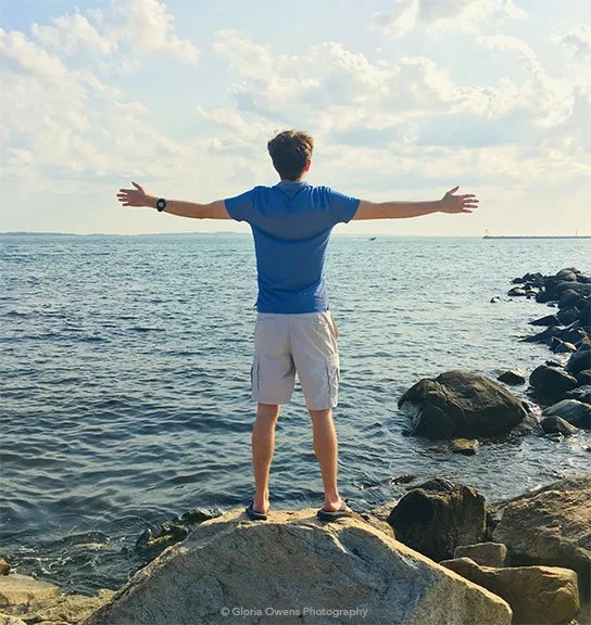A person standing on rocks by the ocean with arms outstretched, facing the water and sky.