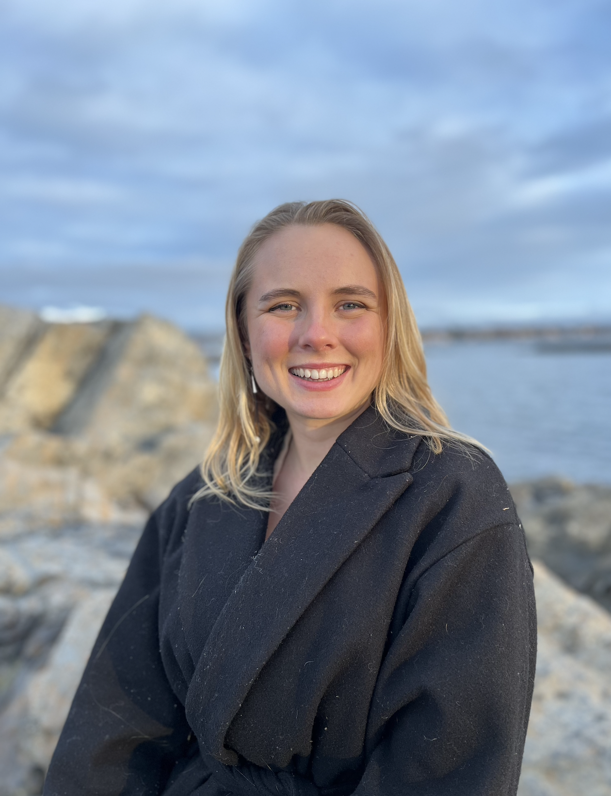A smiling woman with blonde hair standing outdoors near rocks and a body of water with a cloudy sky in the background.