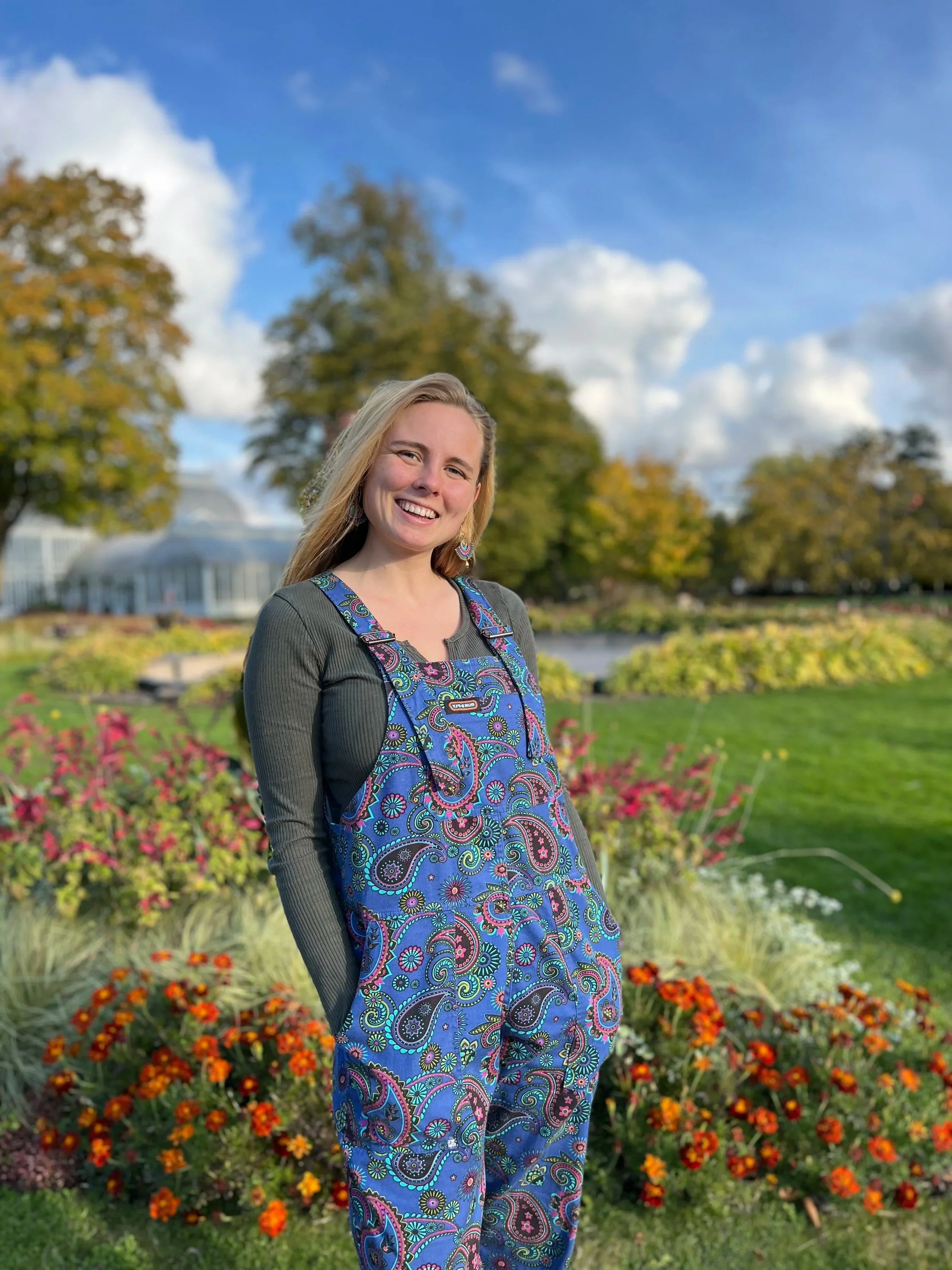 A young woman with blonde hair, smiling, standing outdoors in a park with colorful flowers and trees; she wears a dark long-sleeve shirt and blue paisley overalls.