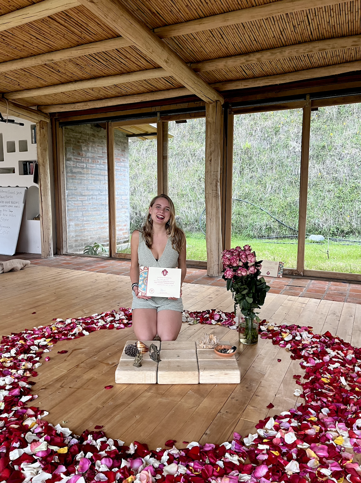 Young woman kneeling inside a heart-shaped border of rose petals, holding a certificate, with a vase of pink roses and small decorative items on a wooden table in front of her, in a rustic indoor setting with large windows and greenery outside.