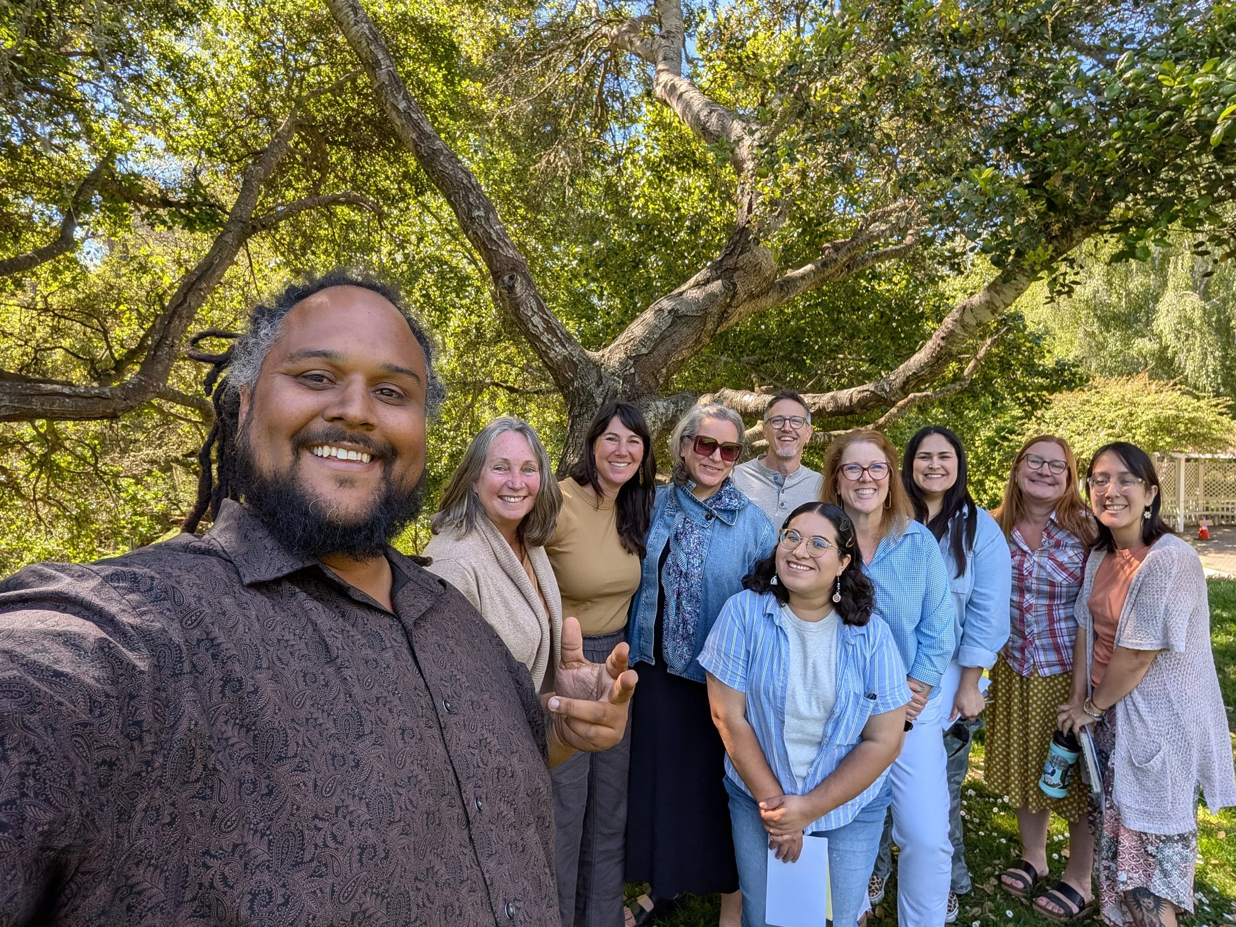 A group of diverse people smiling outdoors in front of a large tree with green leaves.
