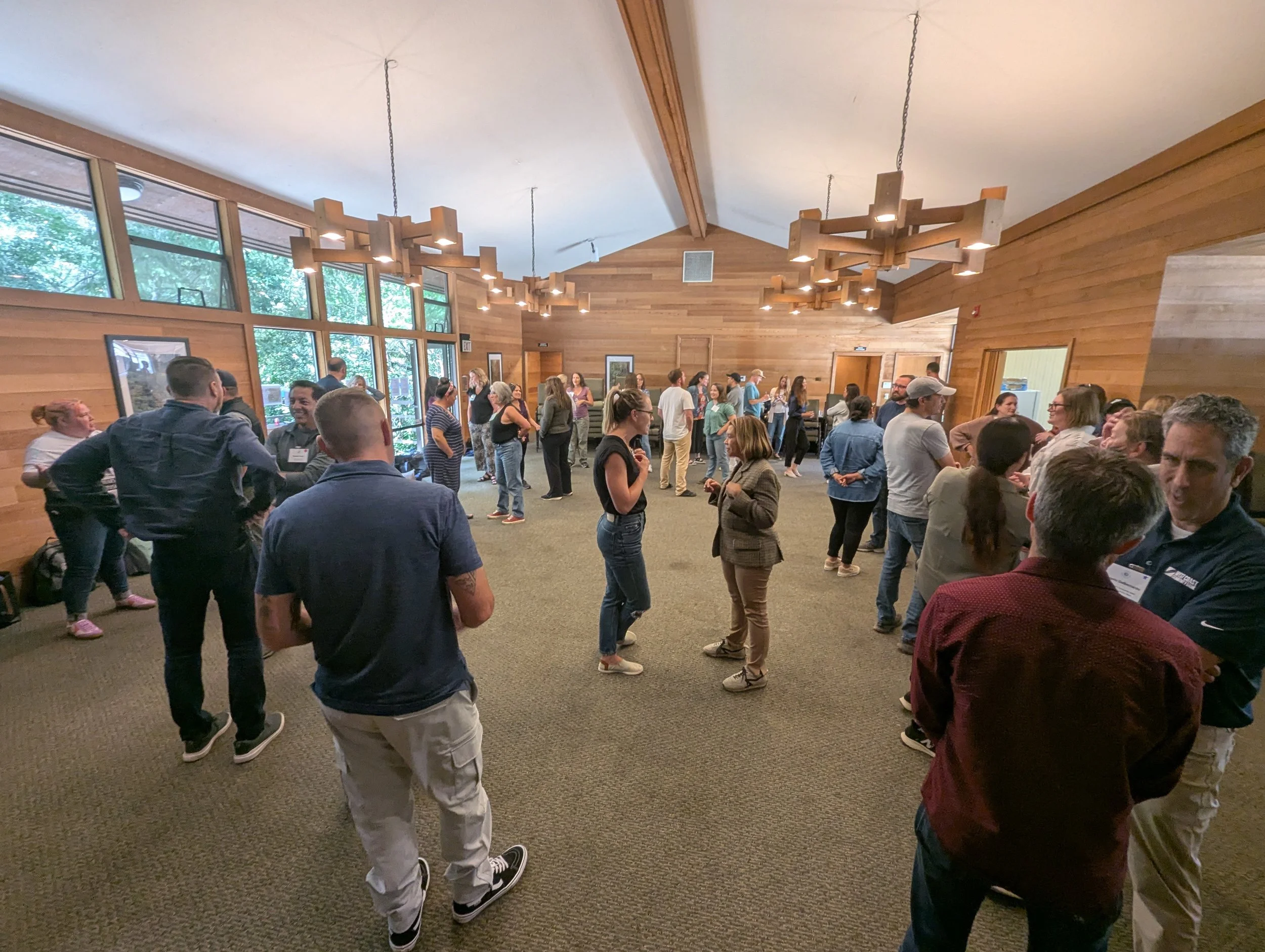 People socializing in a large room with wood-paneled walls and large windows, hanging ceiling lights, and carpeted floor.