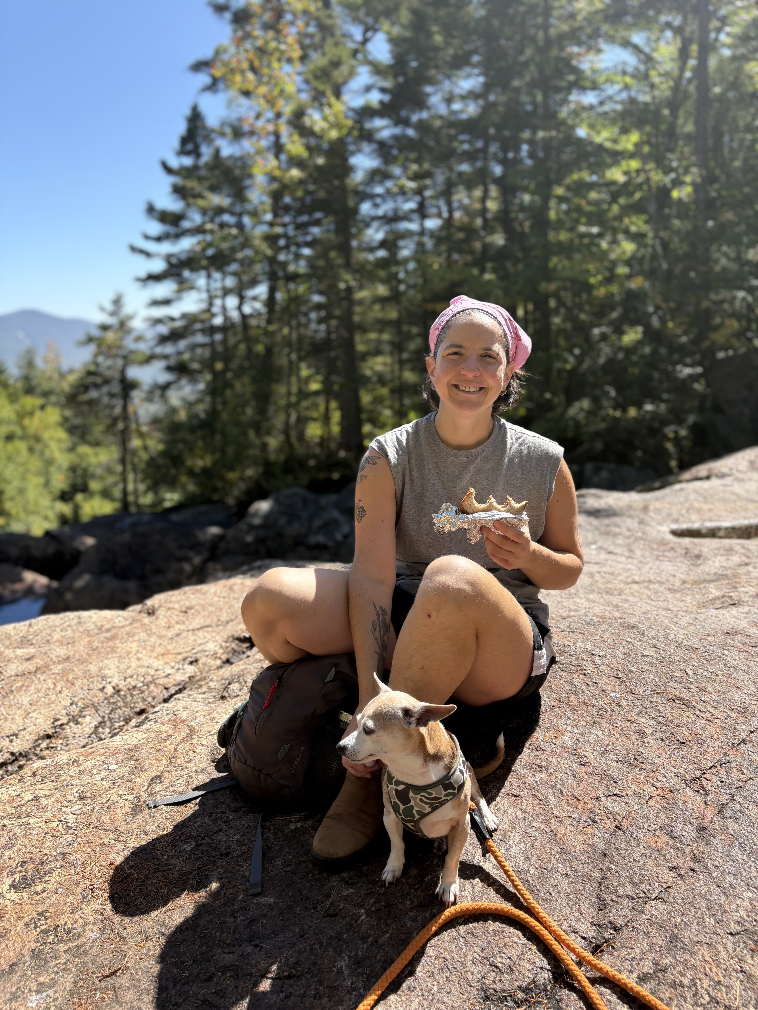 A person smiling, holding a sandwich, and petting their dog on a rock with trees in the background.
