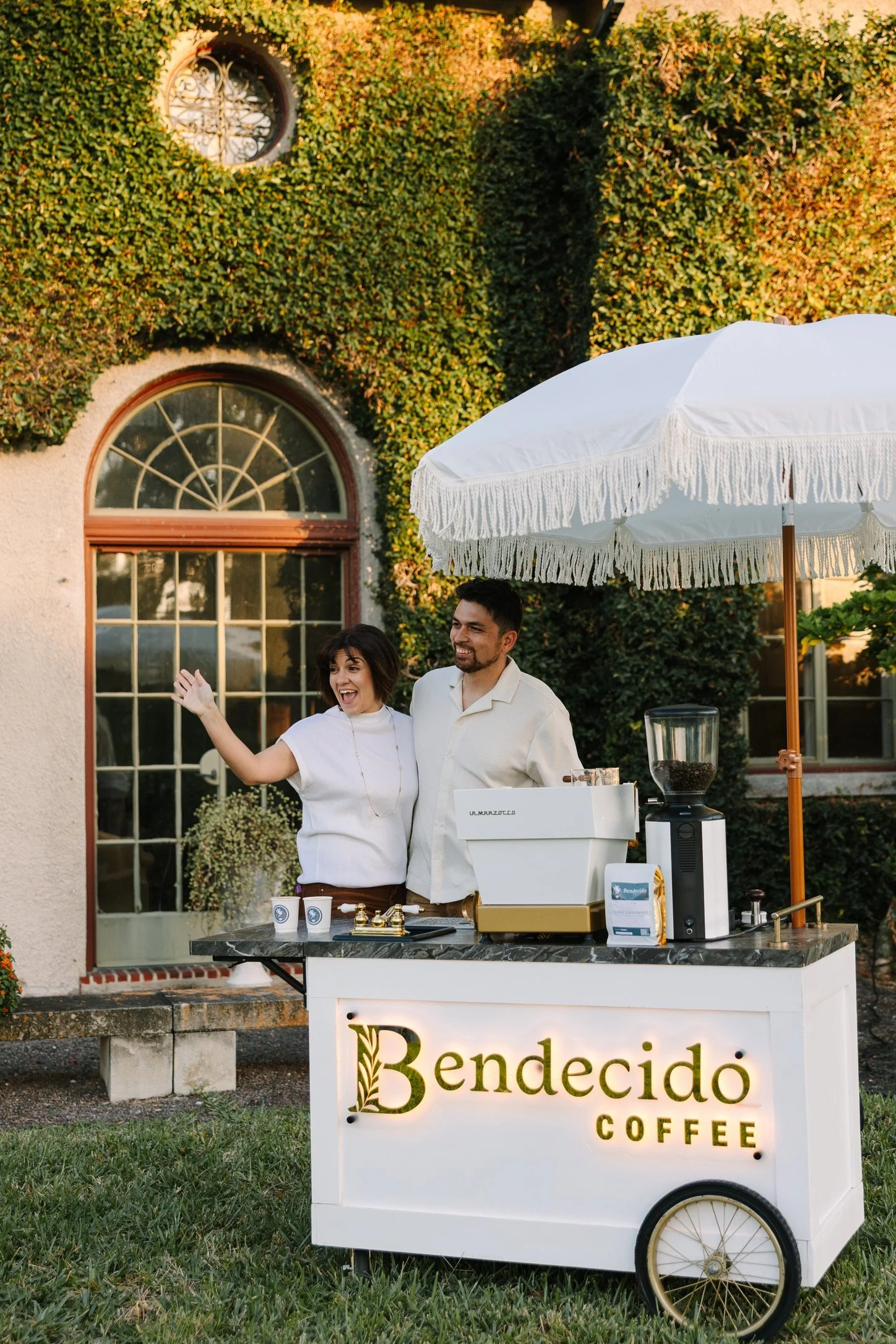 Two Bendecido Coffee baristas working a coffee cart under a white umbrella by an ivy-covered wall