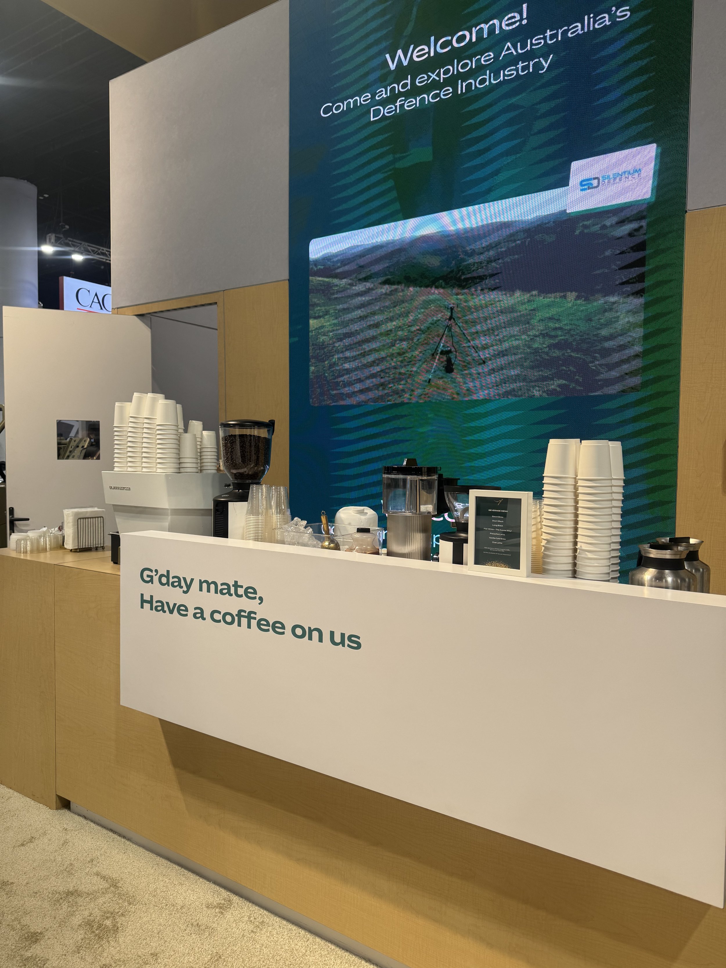 A coffee station at an event with disposable cups, a coffee machine, and various supplies. A large screen behind displays a welcome message and an image of solar panels in a field.