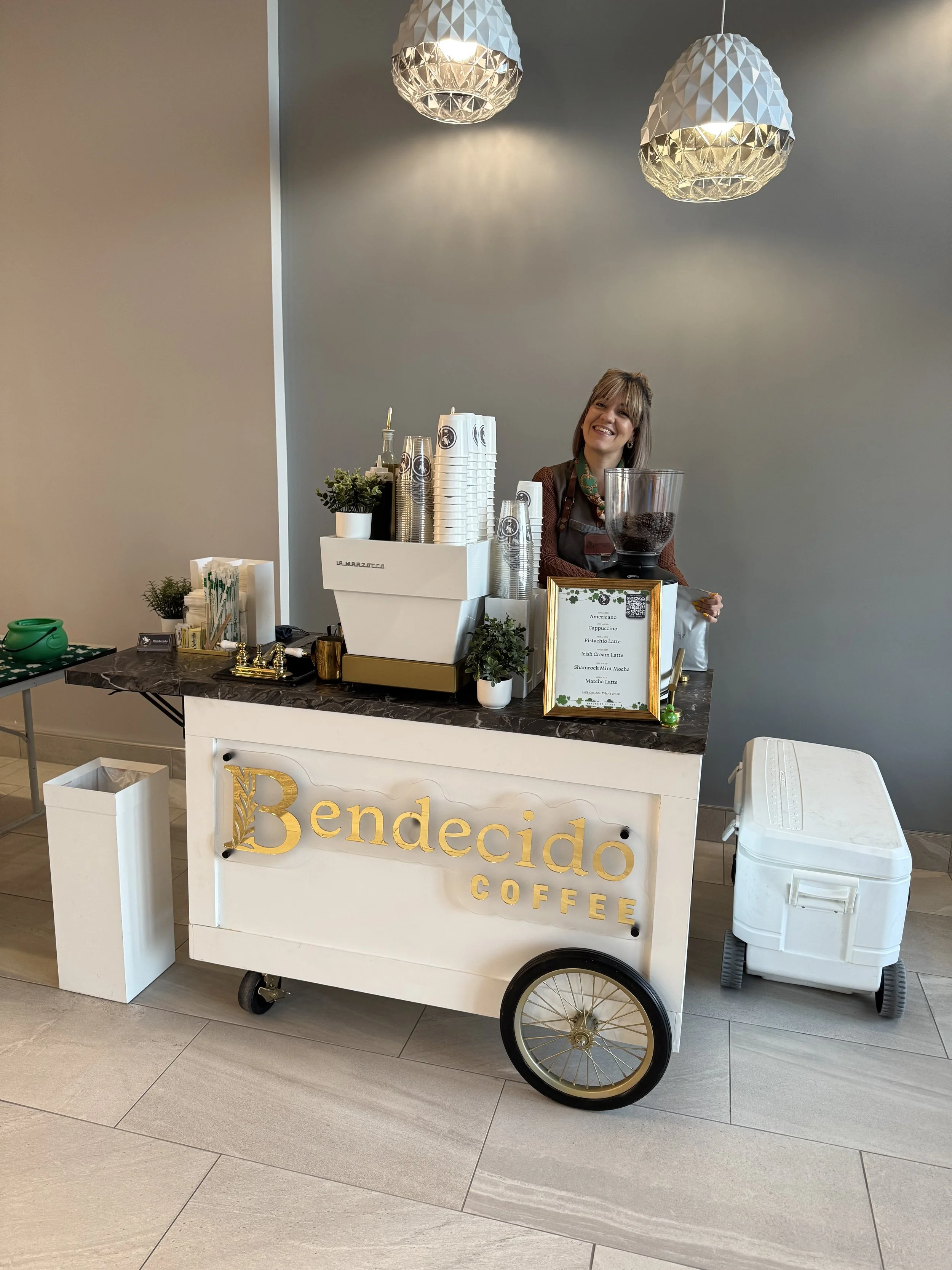 A woman standing behind a white coffee cart with gold lettering that says 'Bendecidó Coffee.' The cart has a black marble top, and there are small potted plants and coffee supplies on it. She is smiling, wearing a necklace, and has a large clear cont