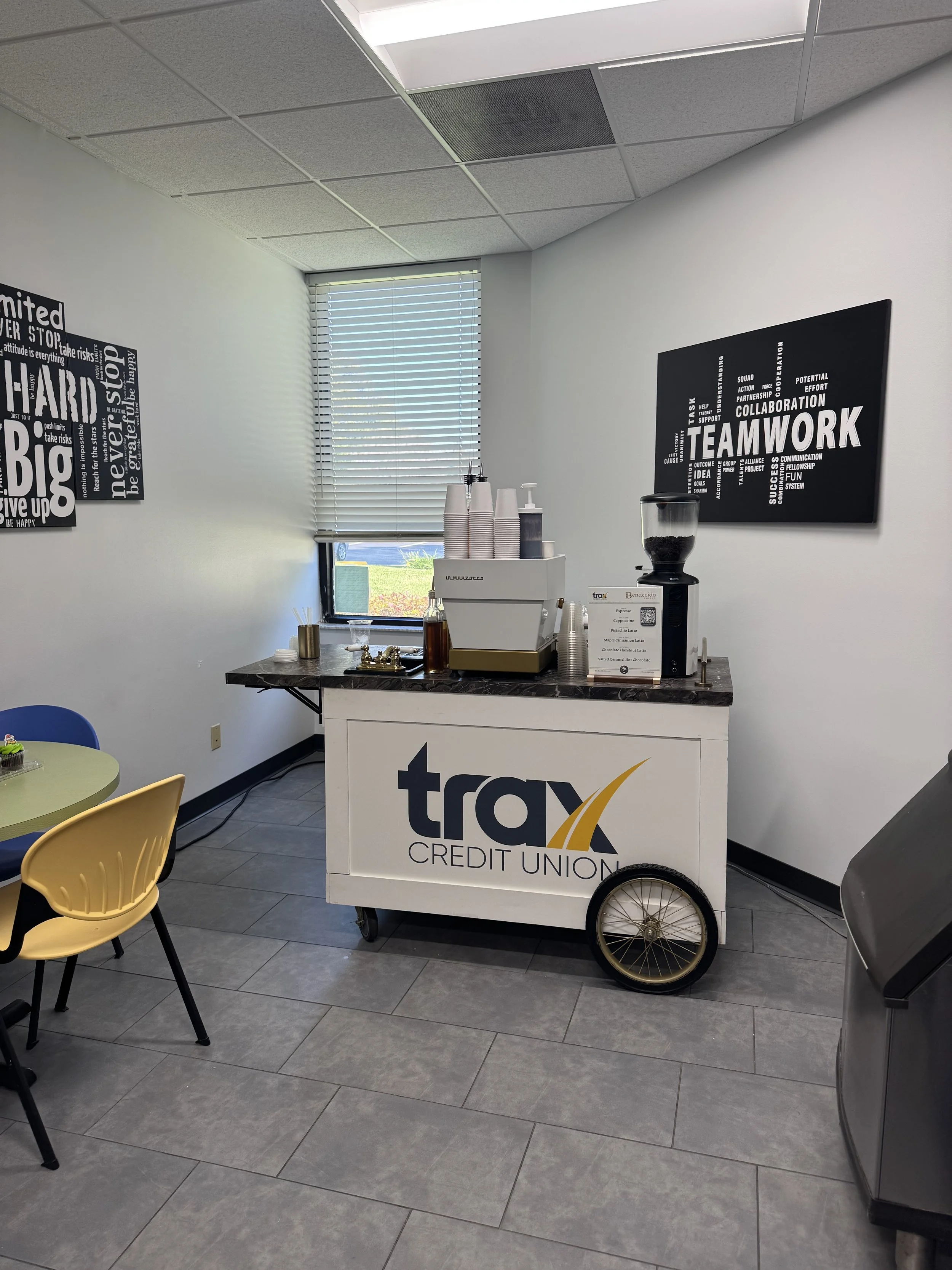 Troy Credit Union beverage station with coffee machine, cups, and decorations, located inside an office or service area. Nearby seating includes a yellow and a blue chair around a small round table.