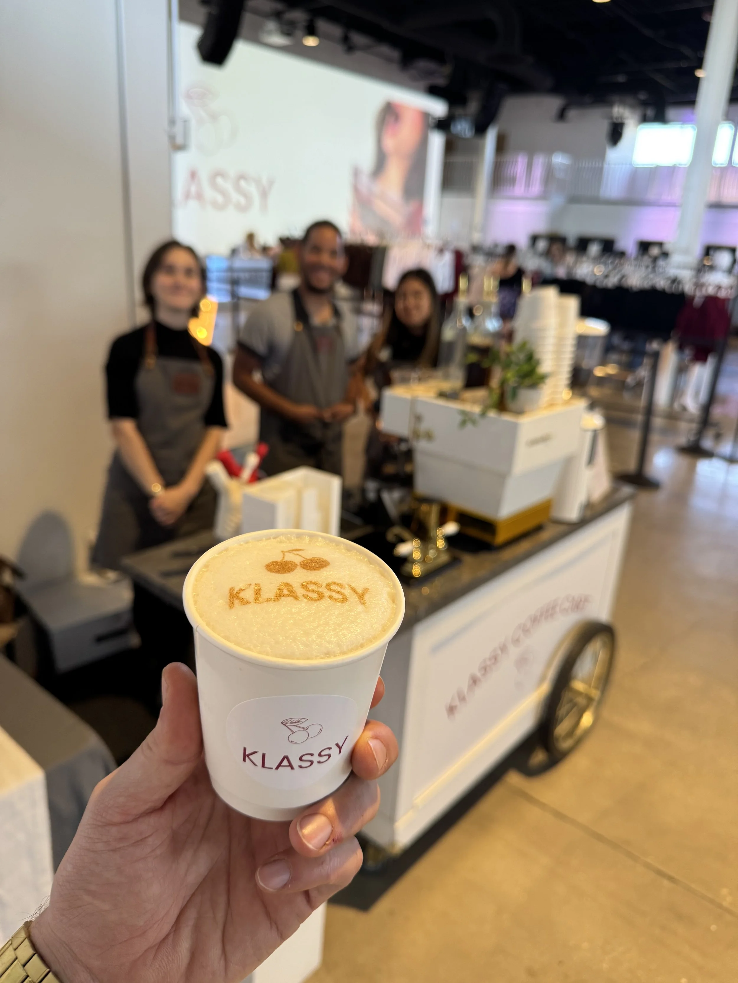 Hand holding a cup of coffee with the word 'KLASSY' written on foam, in front of a coffee cart with three smiling staff members at a marketplace or event.