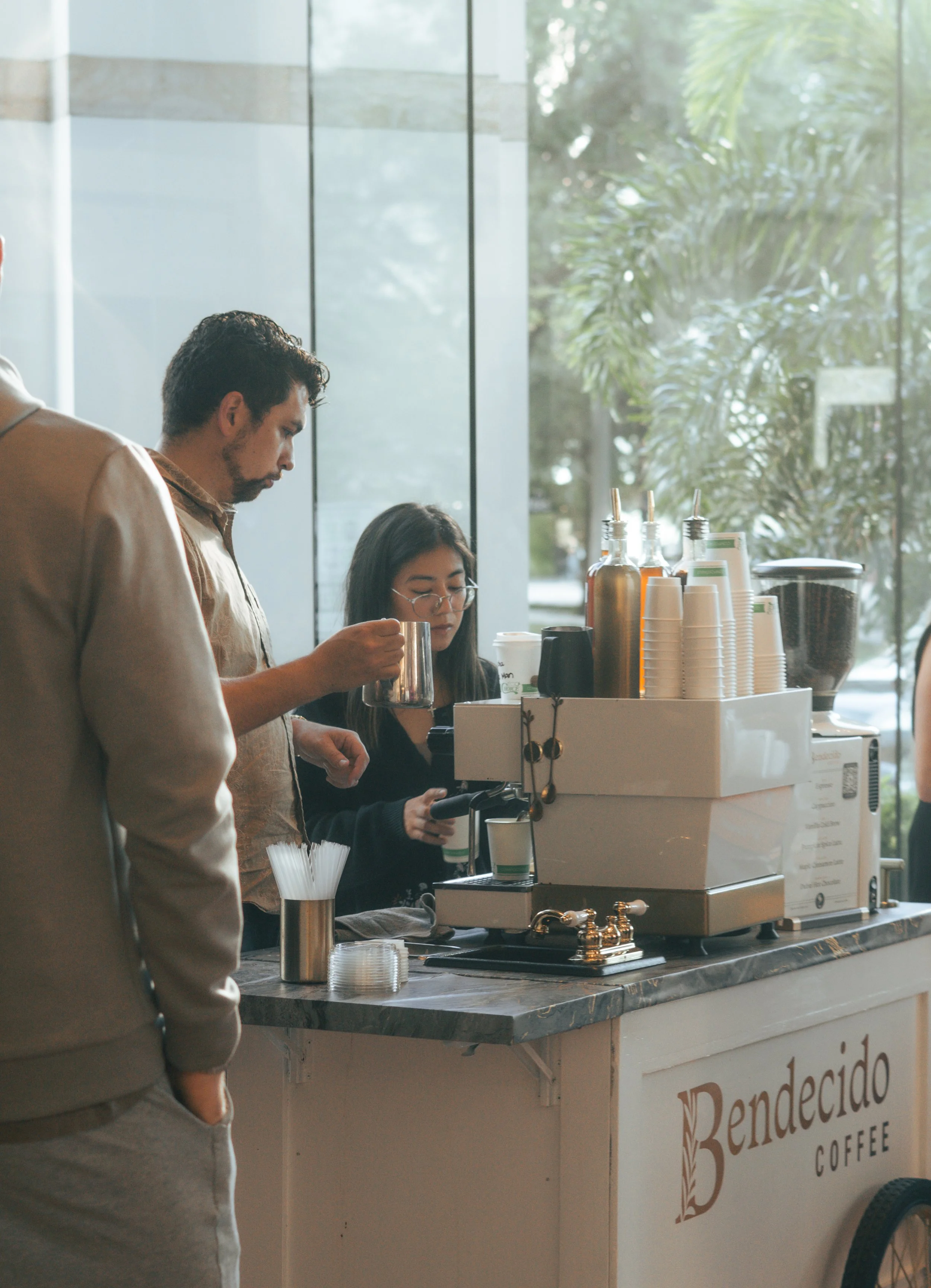 Customers ordering coffee at a cafe counter with a barista preparing a drink, coffee cups, and syrup bottles on display.