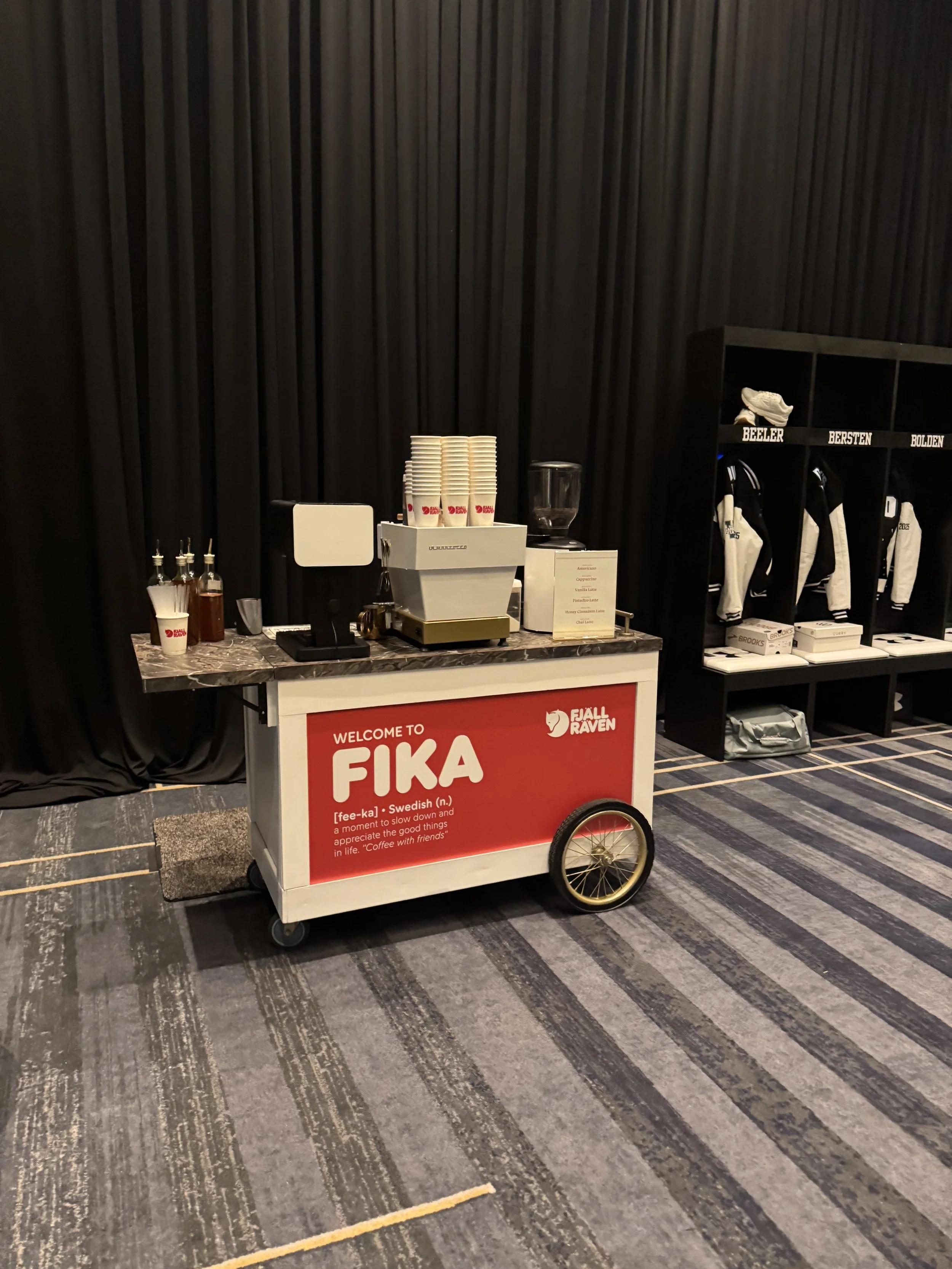 A coffee cart with a red sign reading 'Welcome to FIKA' and a Finnish logo, set against a dark curtain background. The cart has cups, coffee syrup bottles, and equipment for making coffee. Behind the cart, there is a black shelving unit with white an