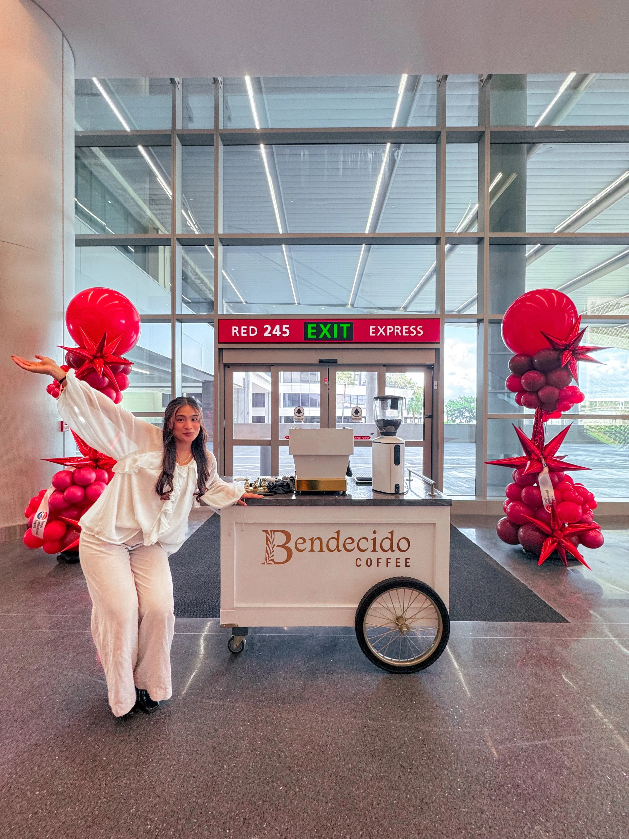 A woman in white clothes posing next to a coffee cart labeled 'Bendecido Coffee' in an indoor area with glass walls, decorated with red balloon arrangements on both sides and a sign above the door displaying 'RED 245 EXIT EXPRESS'.