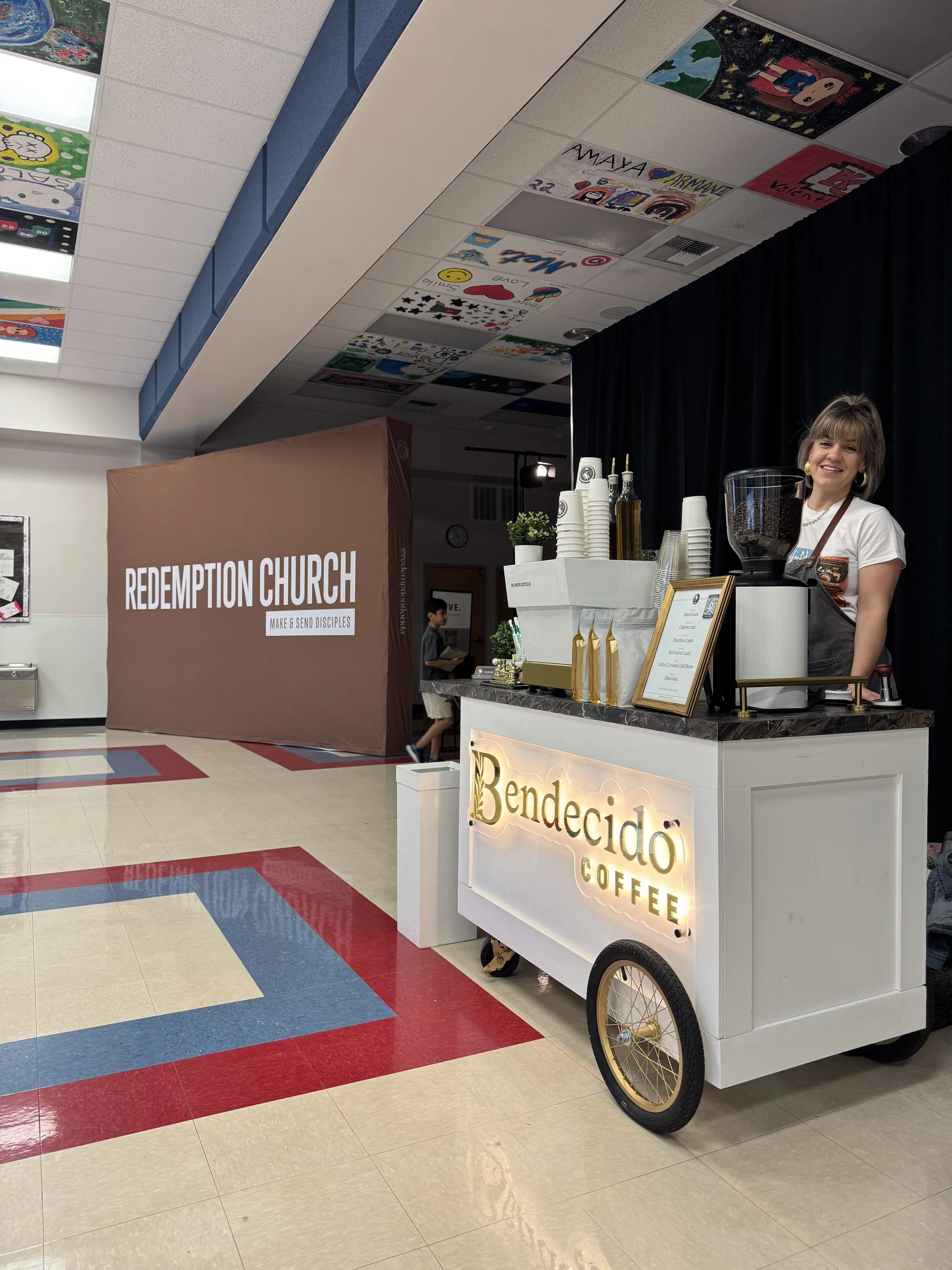 Coffee cart branded with Bendecio Coffee logo served by a smiling woman in a school or community center with colorful ceiling artwork and a brown partition with 'Redemption Church'  of Wesley Chapel written on it.
