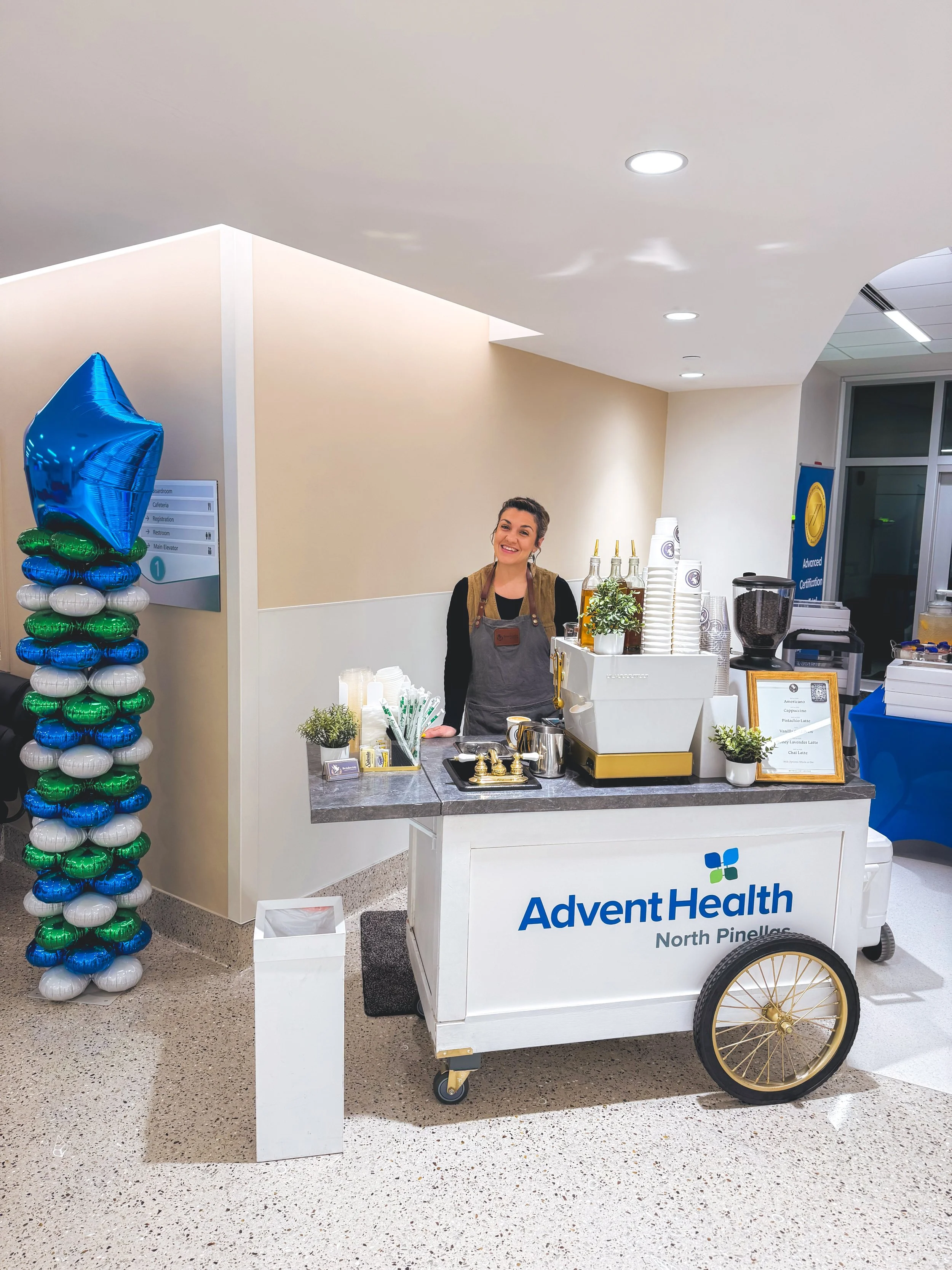 A smiling woman standing at an Advent Health North Pinellas coffee and beverage cart in a hospital lobby, with a blue and green balloon decoration nearby.