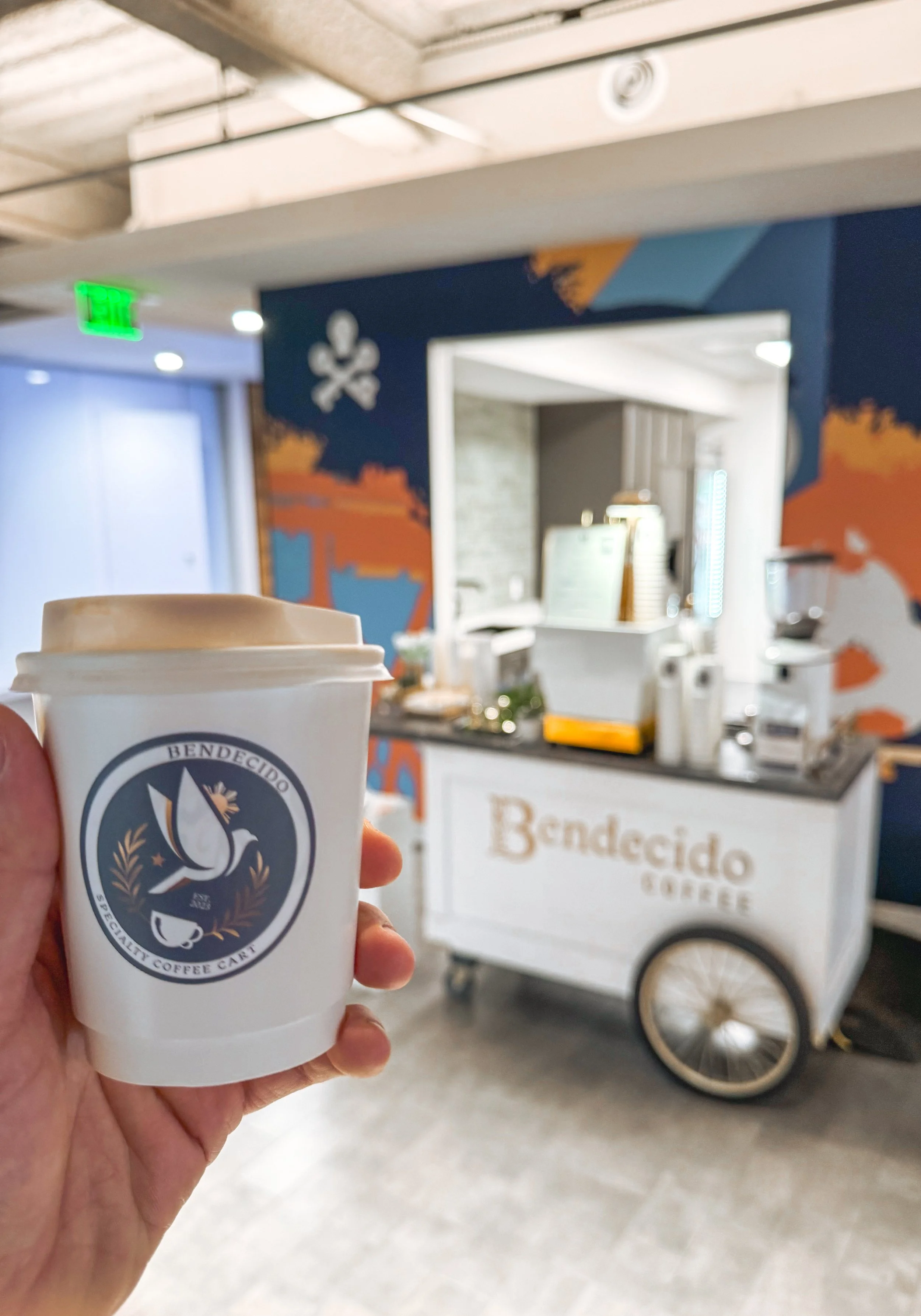 A hand holding a white coffee cup with a blue and white logo in front of a coffee stand that has a sign reading 'Bendecido Coffee' and a colorful Visit Tampa Bay mural on the wall.