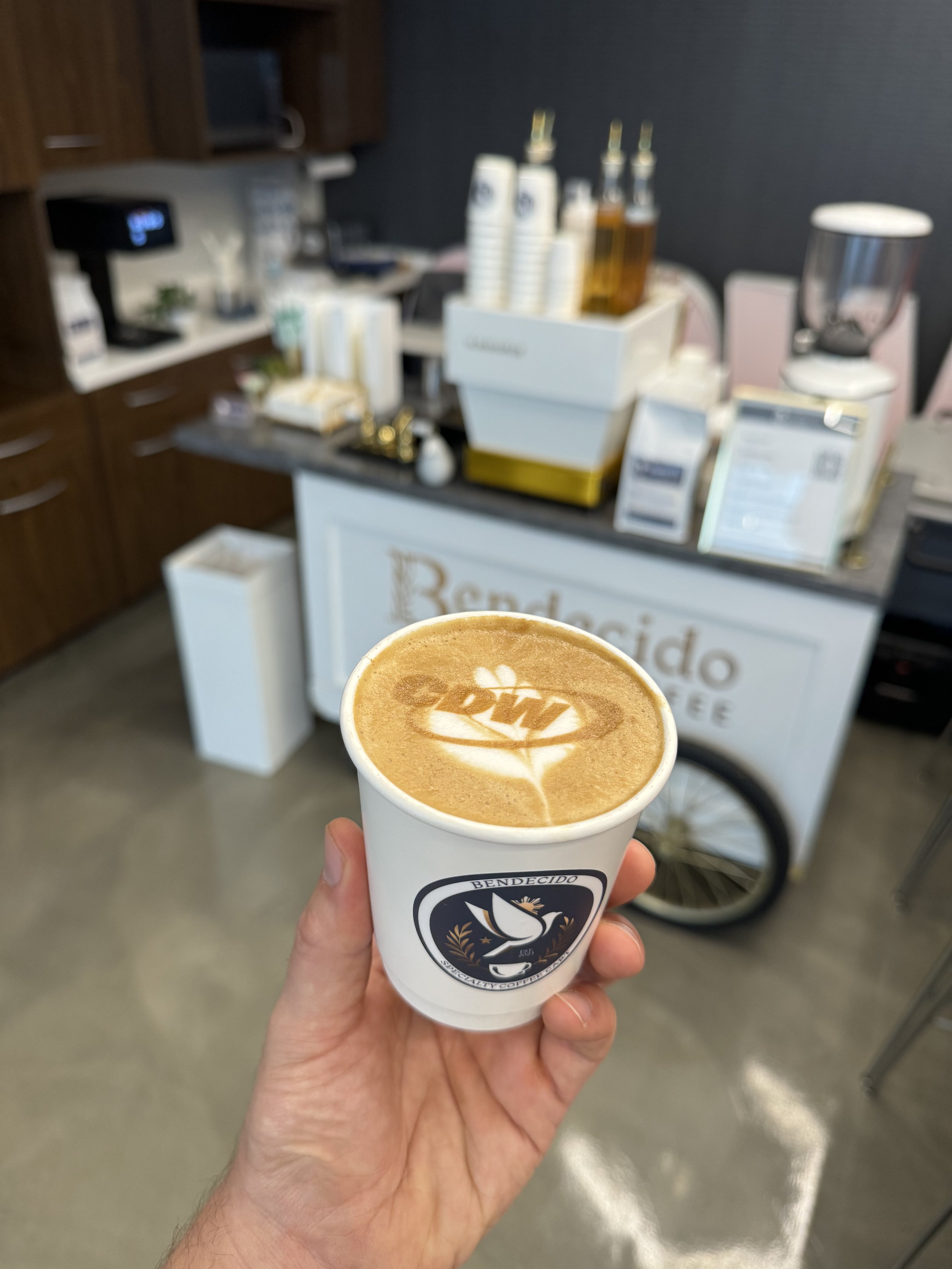 Hand holding a cup of cappuccino with latte art, in front of a coffee cart setup with a logo "Bendecido Coffee" and various equipment in a cafe setting.