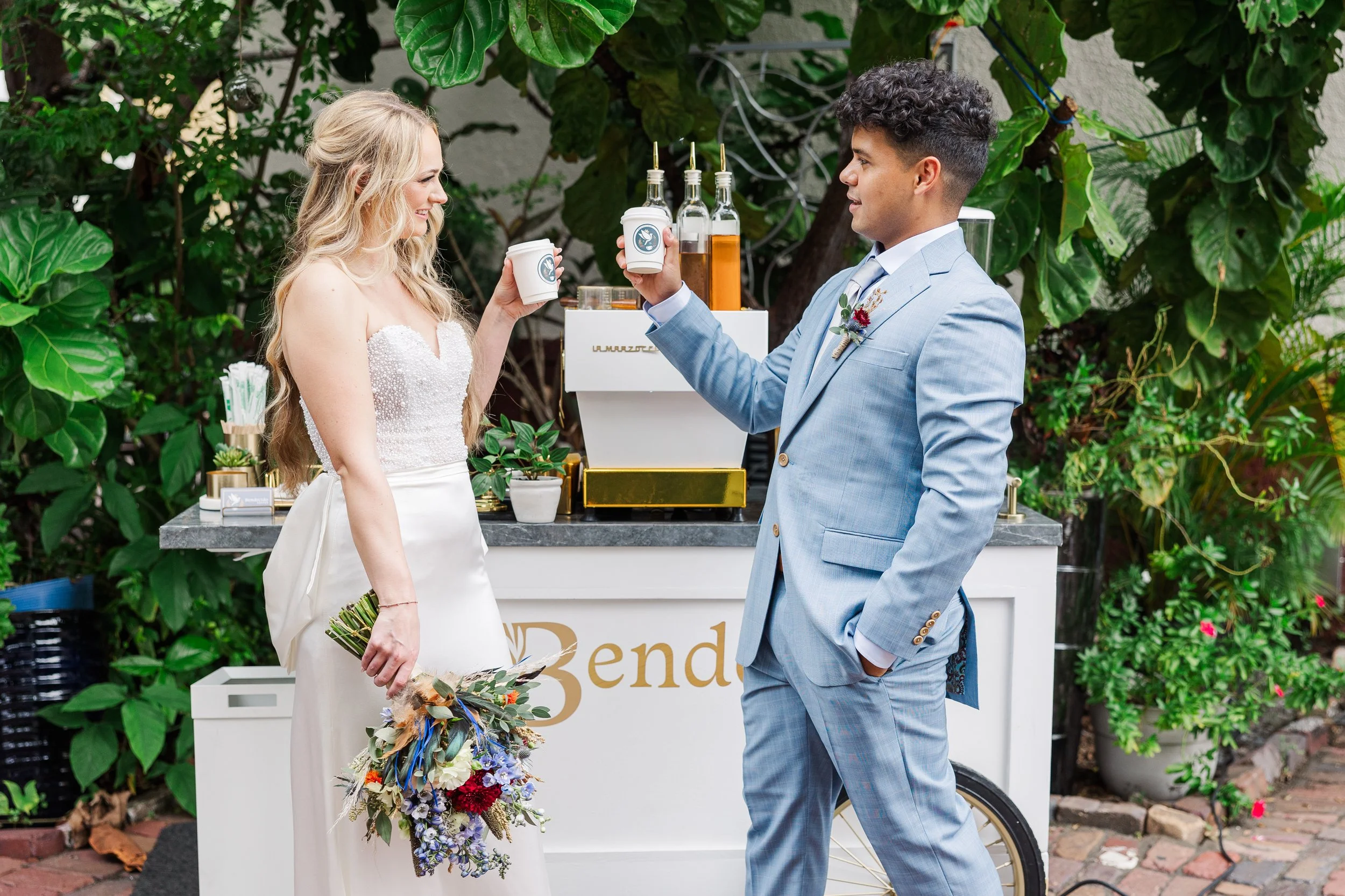 A couple having a toast at an outdoor wedding reception, with the bride holding a bouquet and the groom wearing a light blue suit, standing in front of a drink cart with greenery in the background.
