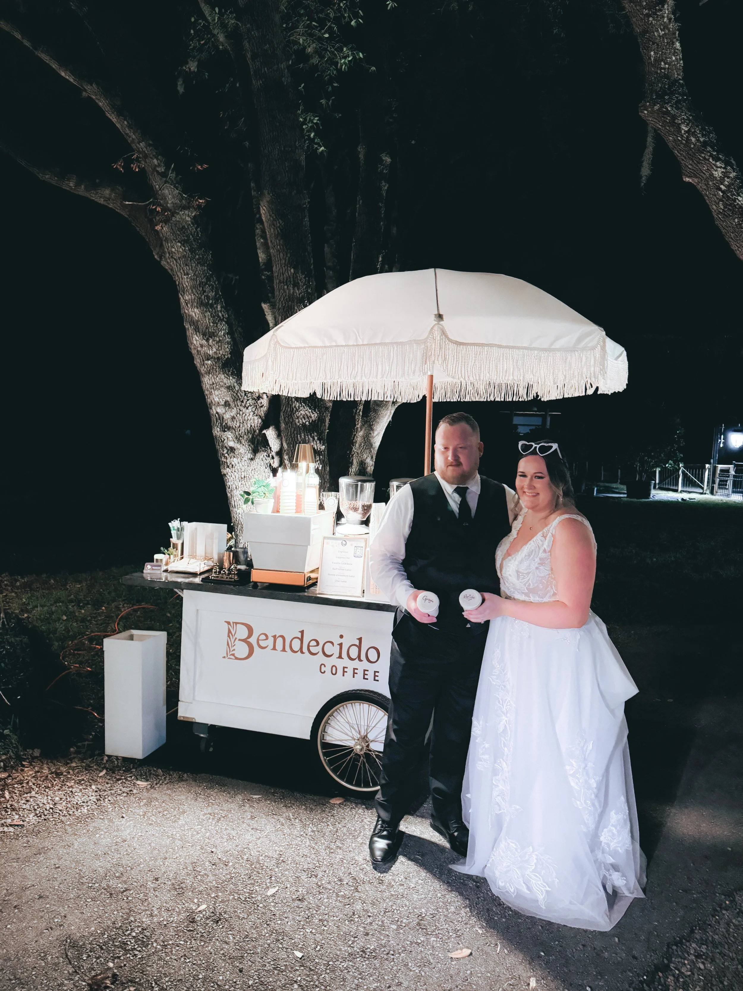 A newlywed couple standing next to a coffee cart under a large tree at night, with the bride in a white wedding dress and the groom in a black vest and tie, both holding coffee cups, with the cart labeled 'Bendecido Coffee'.