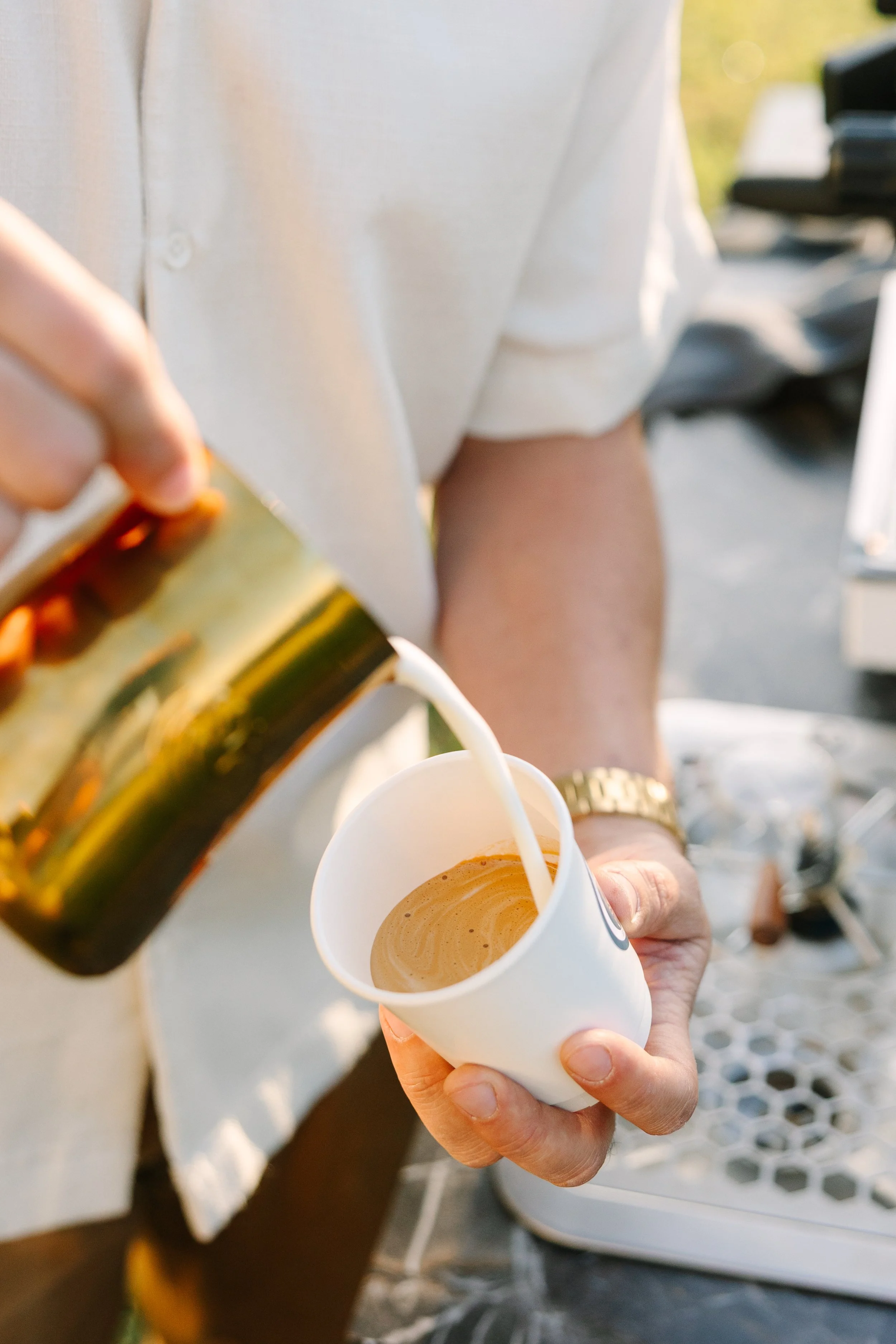 A person in a white shirt pouring milk into a cup of coffee at an outdoor setting.