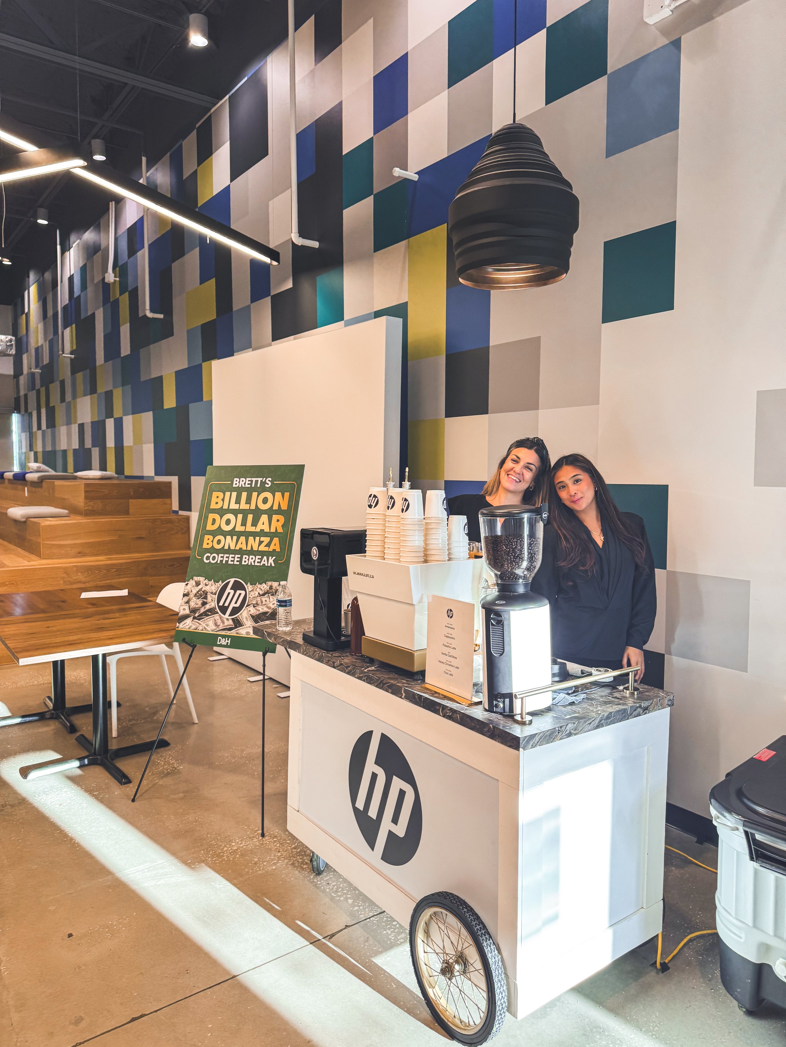 Two women standing behind a coffee cart with an HP logo, inside a modern cafe with geometric patterned wall and wooden tables.