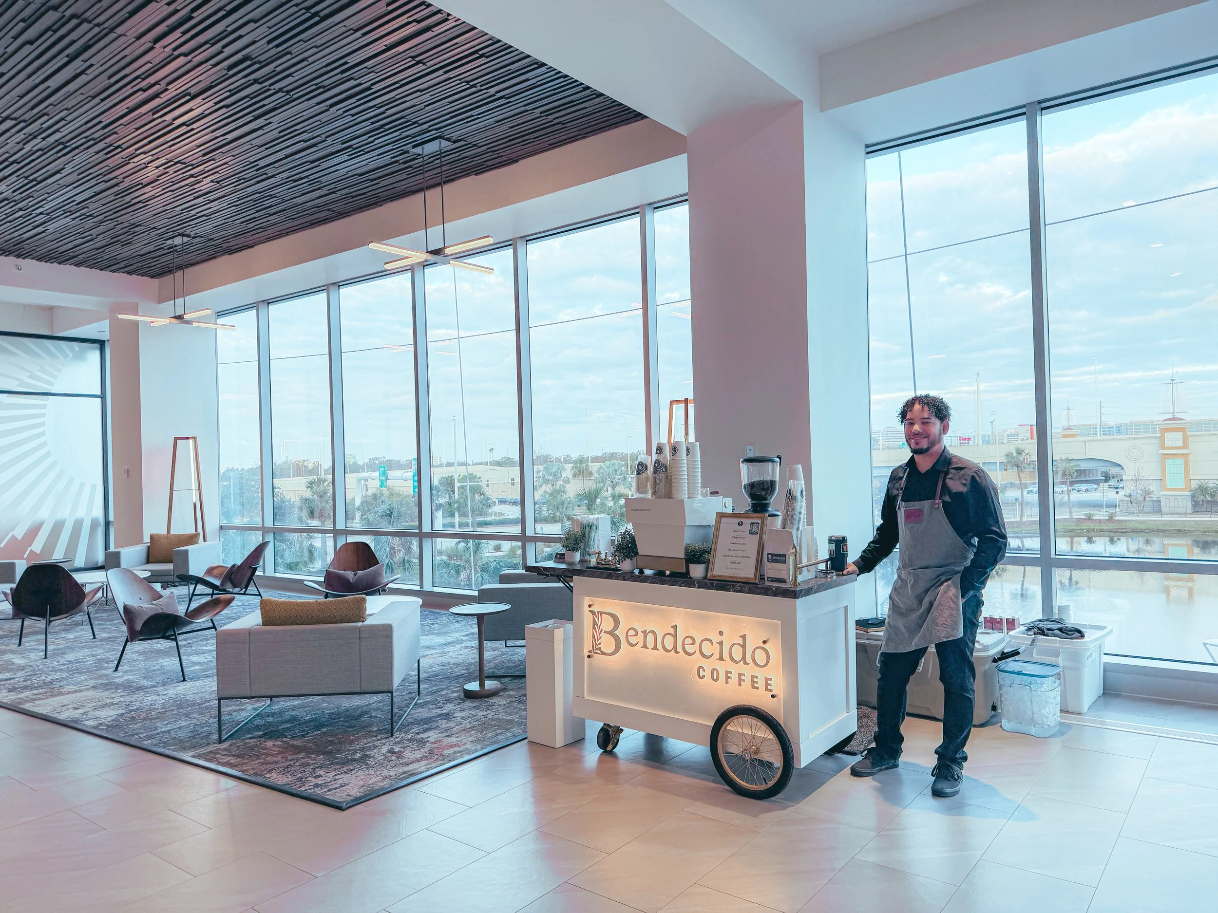 A man in a black shirt and gray apron standing by a coffee cart labeled "Bendecido Coffee" inside a modern, spacious lounge with large windows, seating area, and city view from Midtown Tampa.