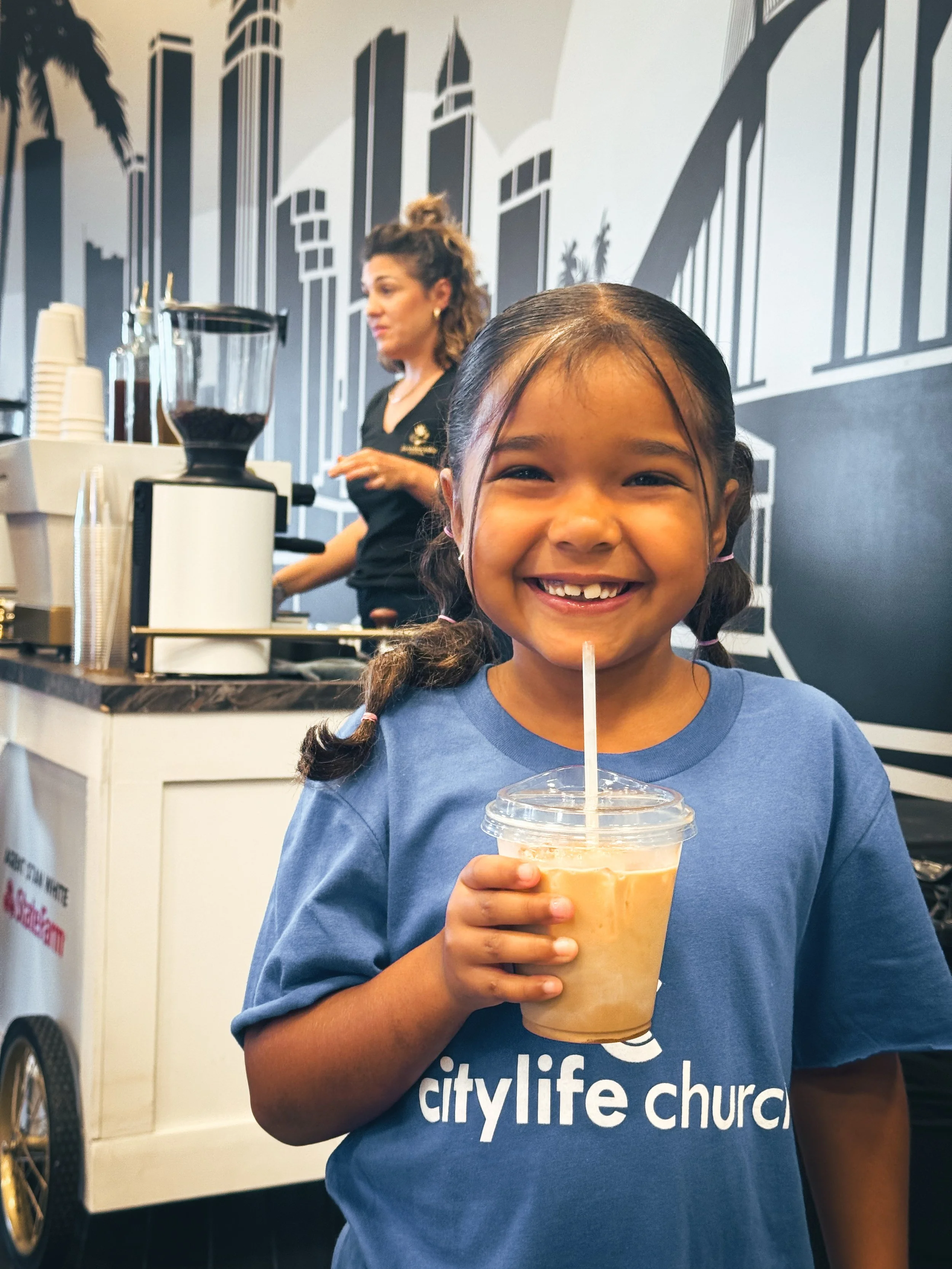Smiling young girl holding a cup of iced coffee in a cafe with a barista in the background and a cityscape mural on the wall.