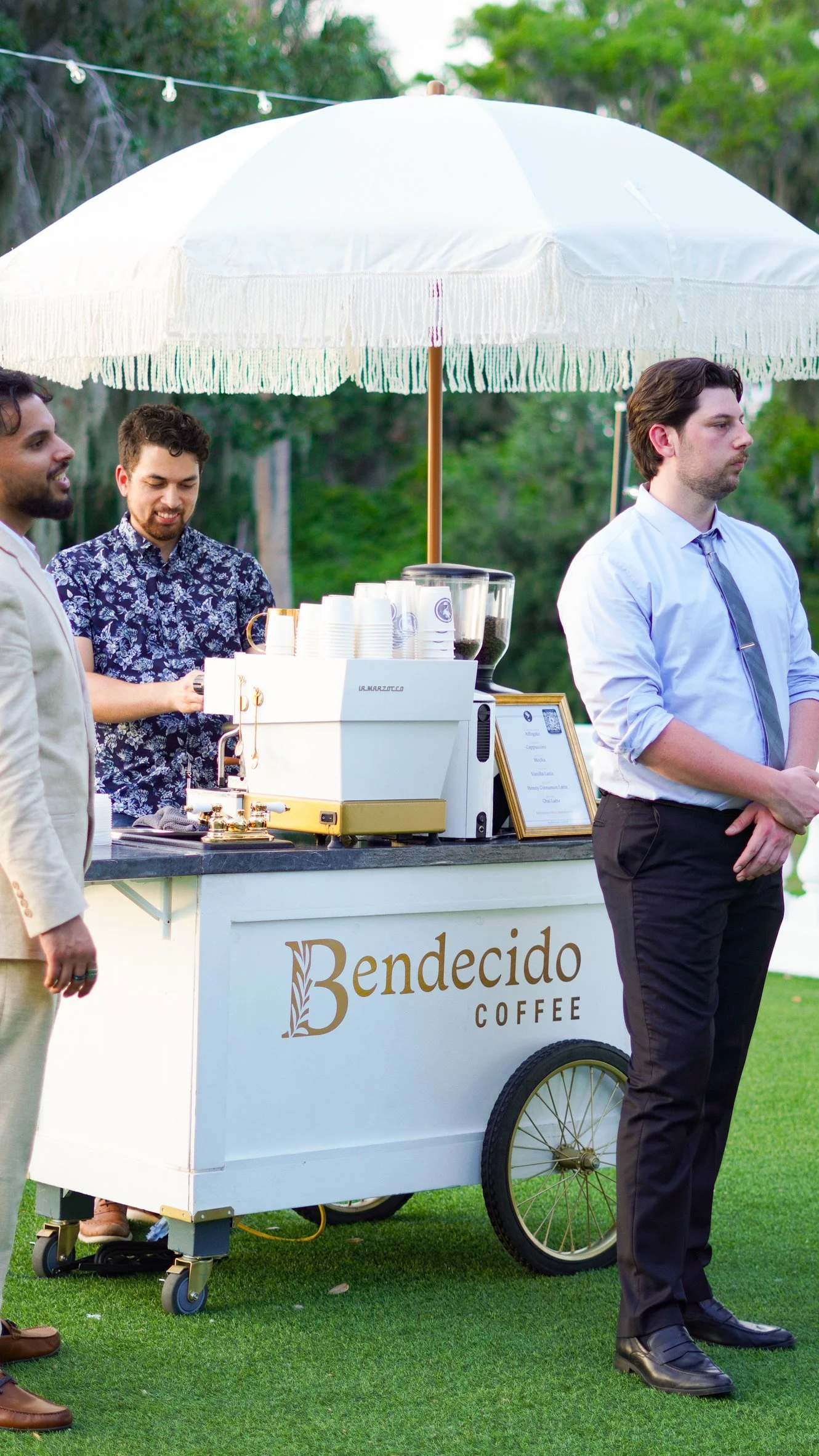Two men standing beside a Bendecido Coffee cart under a white umbrella on a green lawn