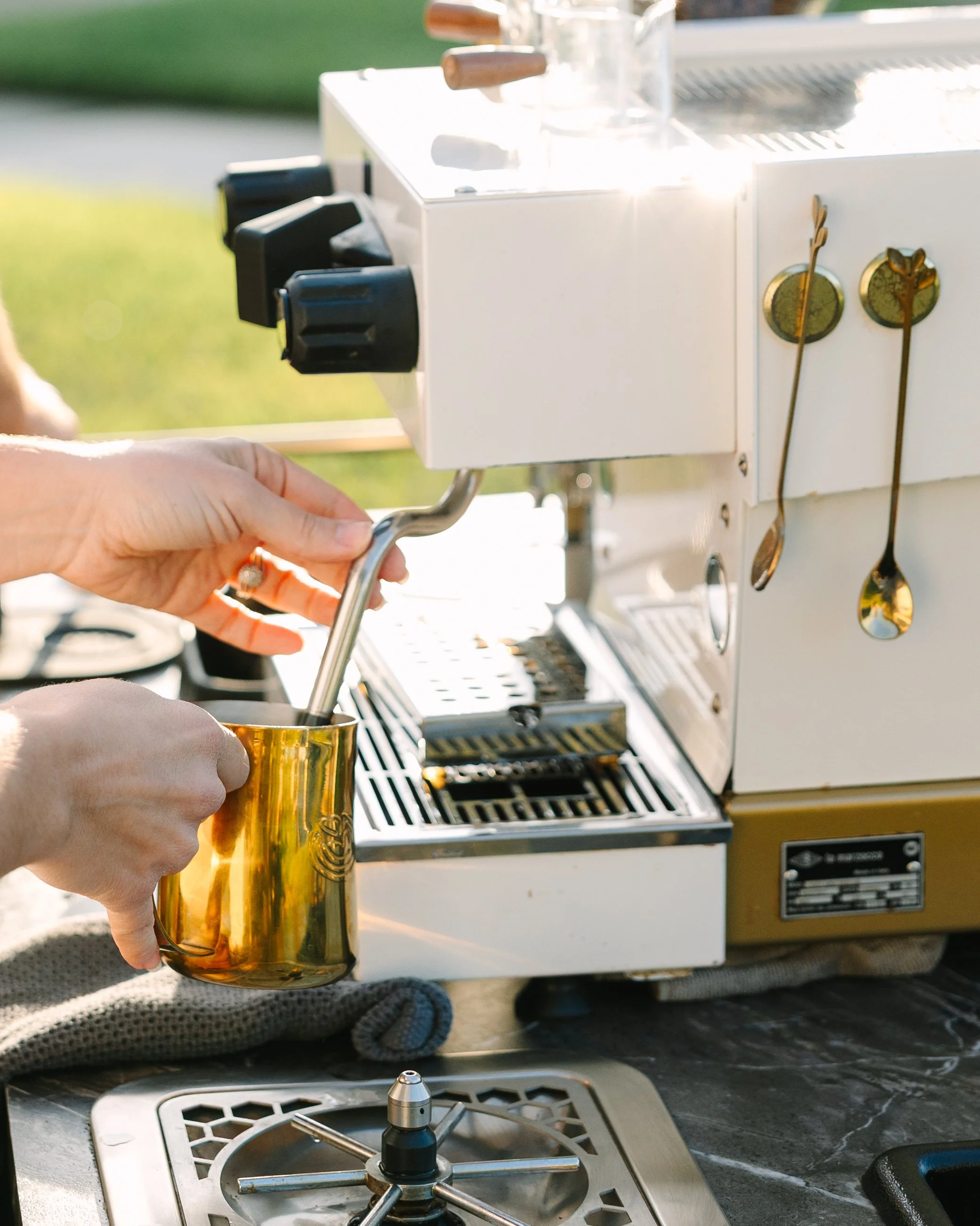 Person making espresso with a coffee machine outdoors, using a gold-colored metal pitcher.