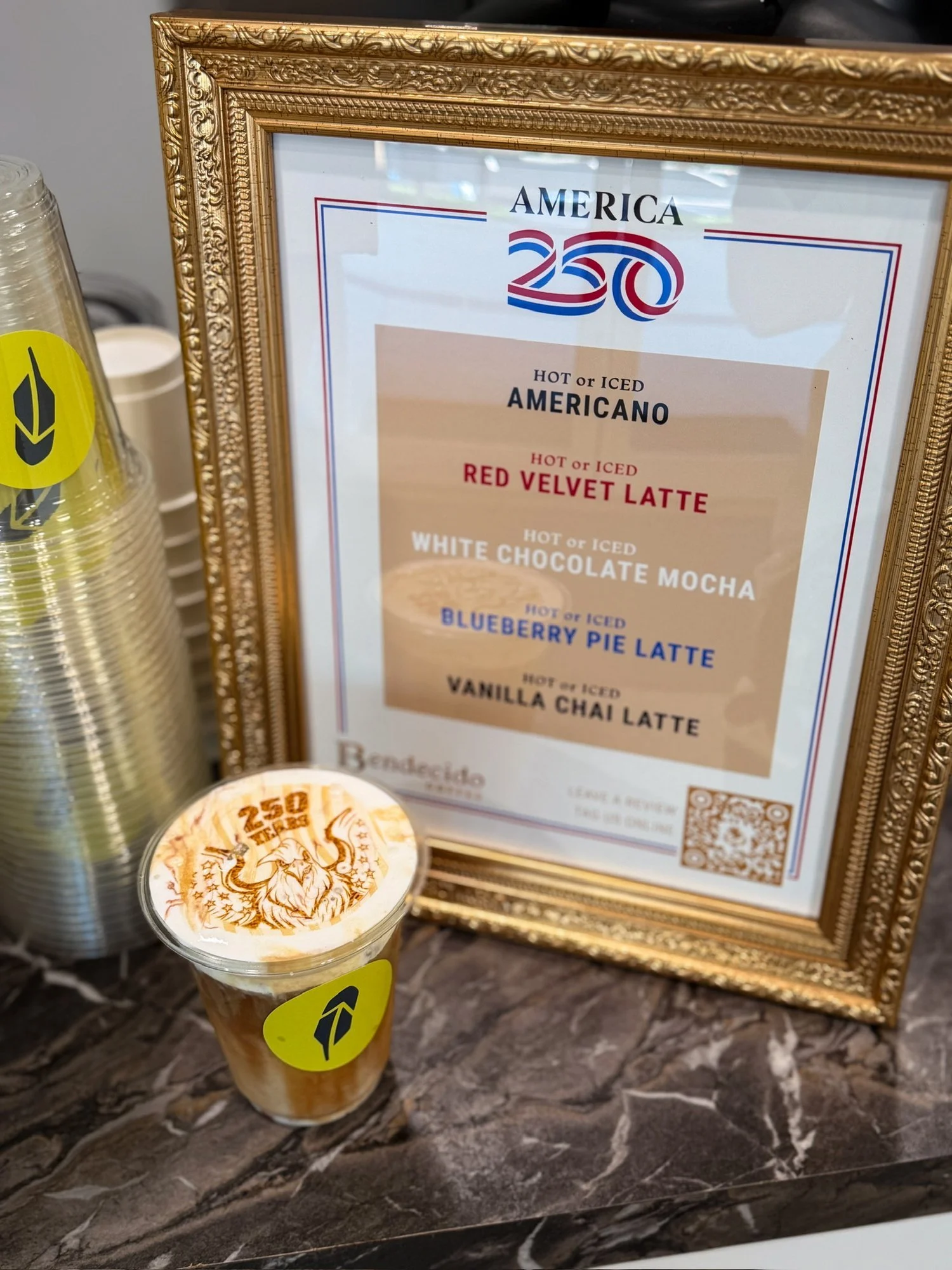 Latte with foam art beside a gold-framed America 250 specialty drink menu from Bendecido Coffee