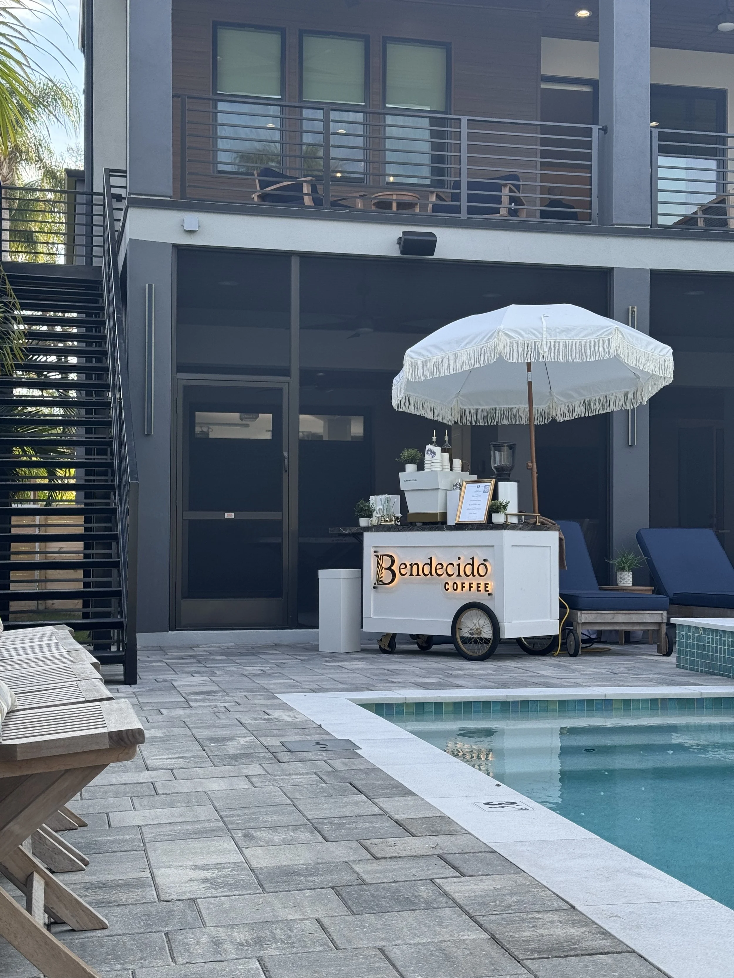 A coffee cart labeled 'Bendecido Coffee' with a white umbrella, set up by a swimming pool at a modern apartment complex near Downtown Tampa.